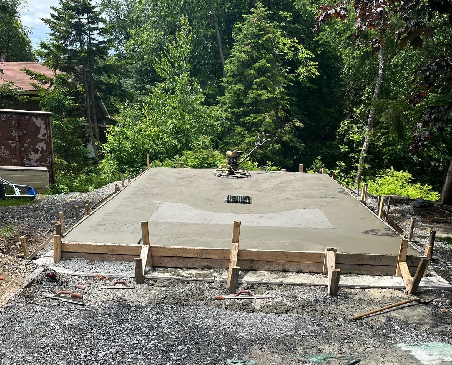Fondations en béton frais coulé dans des coffrages en bois, installées dans une cour gravillonnée, avec des arbres en arrière-plan.