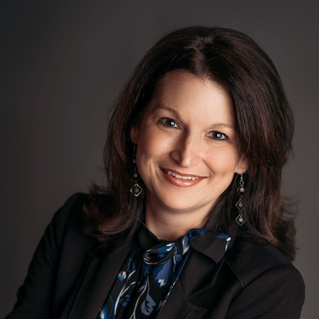Smiling woman with dark hair in a black outfit and blue patterned scarf against a dark background