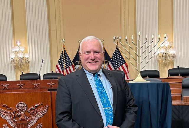 Man in a suit smiling in a formal government chamber with U.S. flags behind him