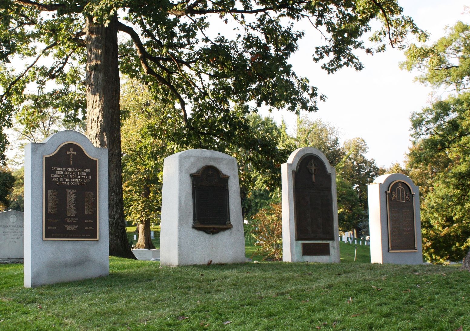 Four gravestones in a sunny cemetery under trees, with grass and dappled light.