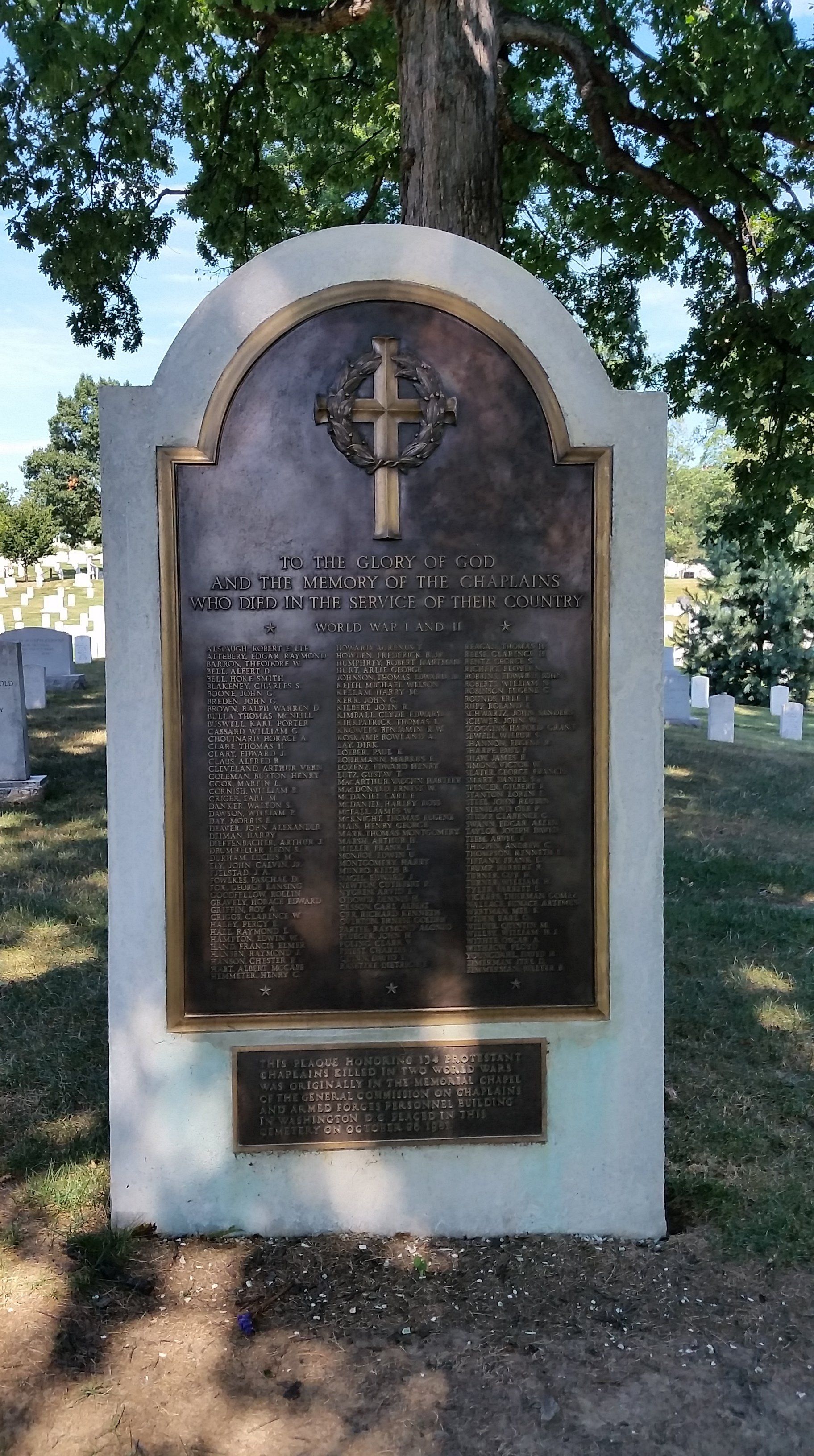 Bronze memorial plaque on a white stone monument in a cemetery, with a cross at the top.