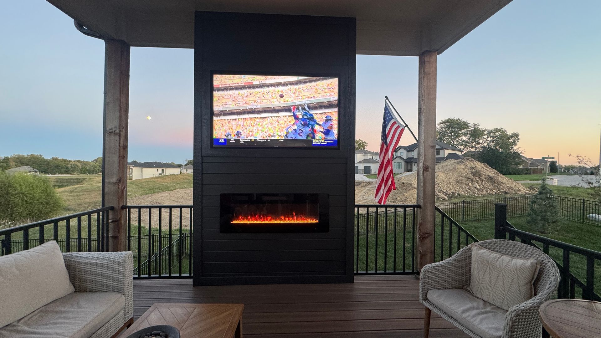 Outdoor patio with TV and fireplace; seating, American flag, and scenic background.