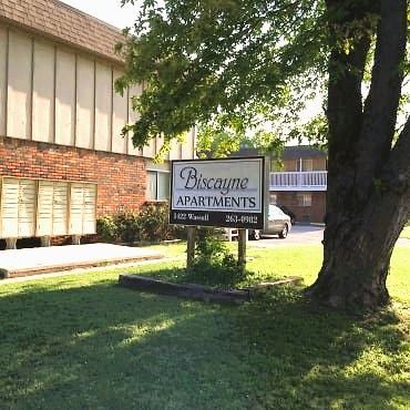 Biscayne Apartments sign in front of a building with brick and beige siding. Green lawn, tree, and parked car are visible.
