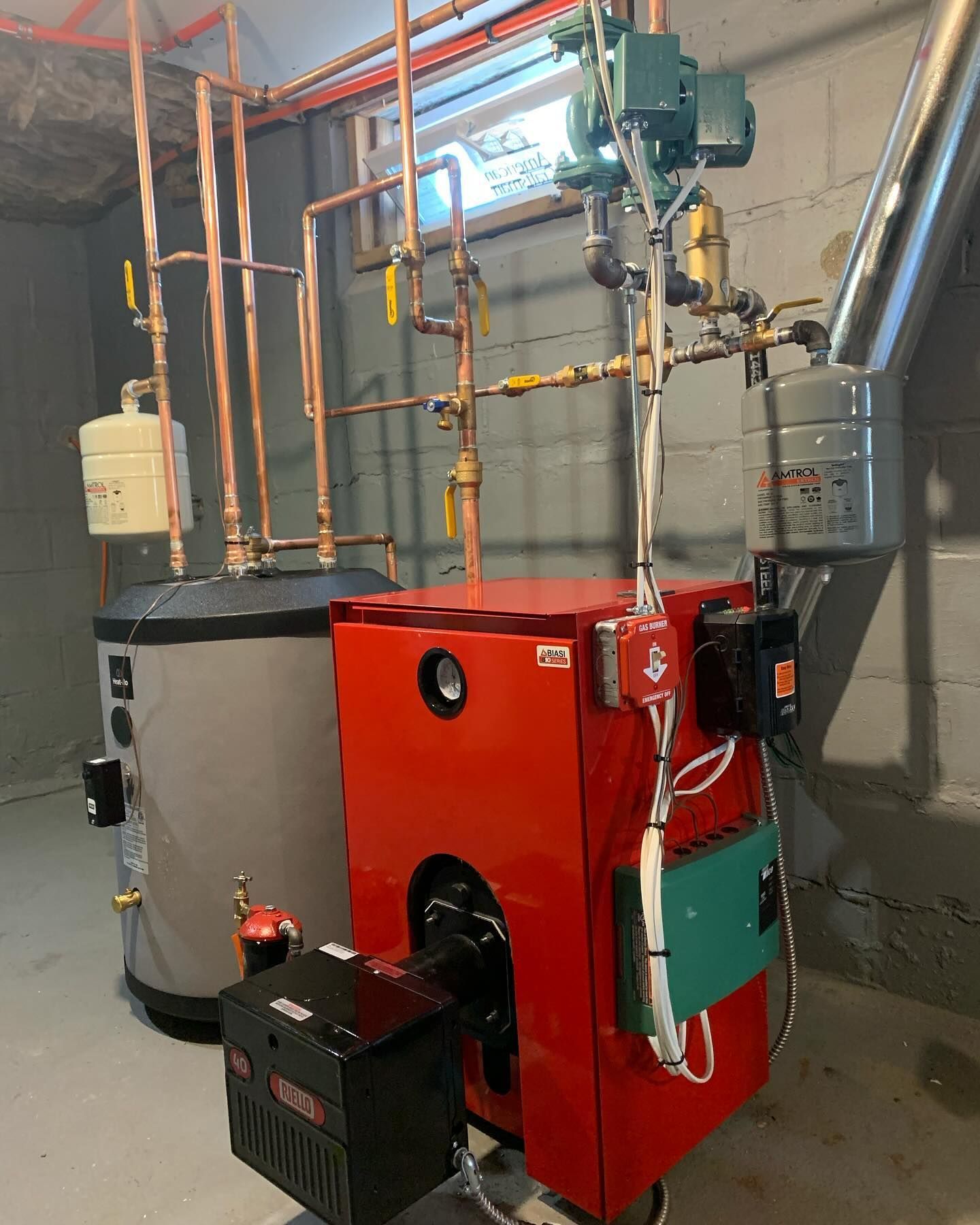 A bright red oil-fired boiler and a gray hot water tank with copper plumbing connections in a basement setting.