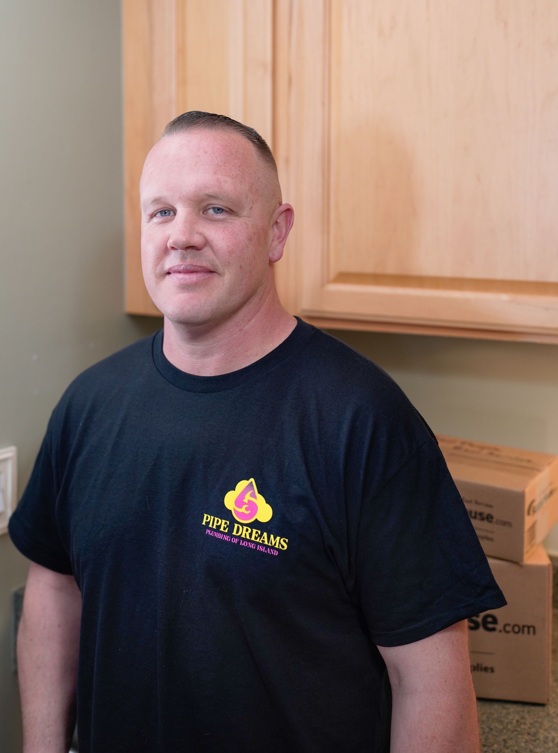 A person with a short haircut wearing a black t-shirt with a logo, standing indoors in front of light-wood cabinets.