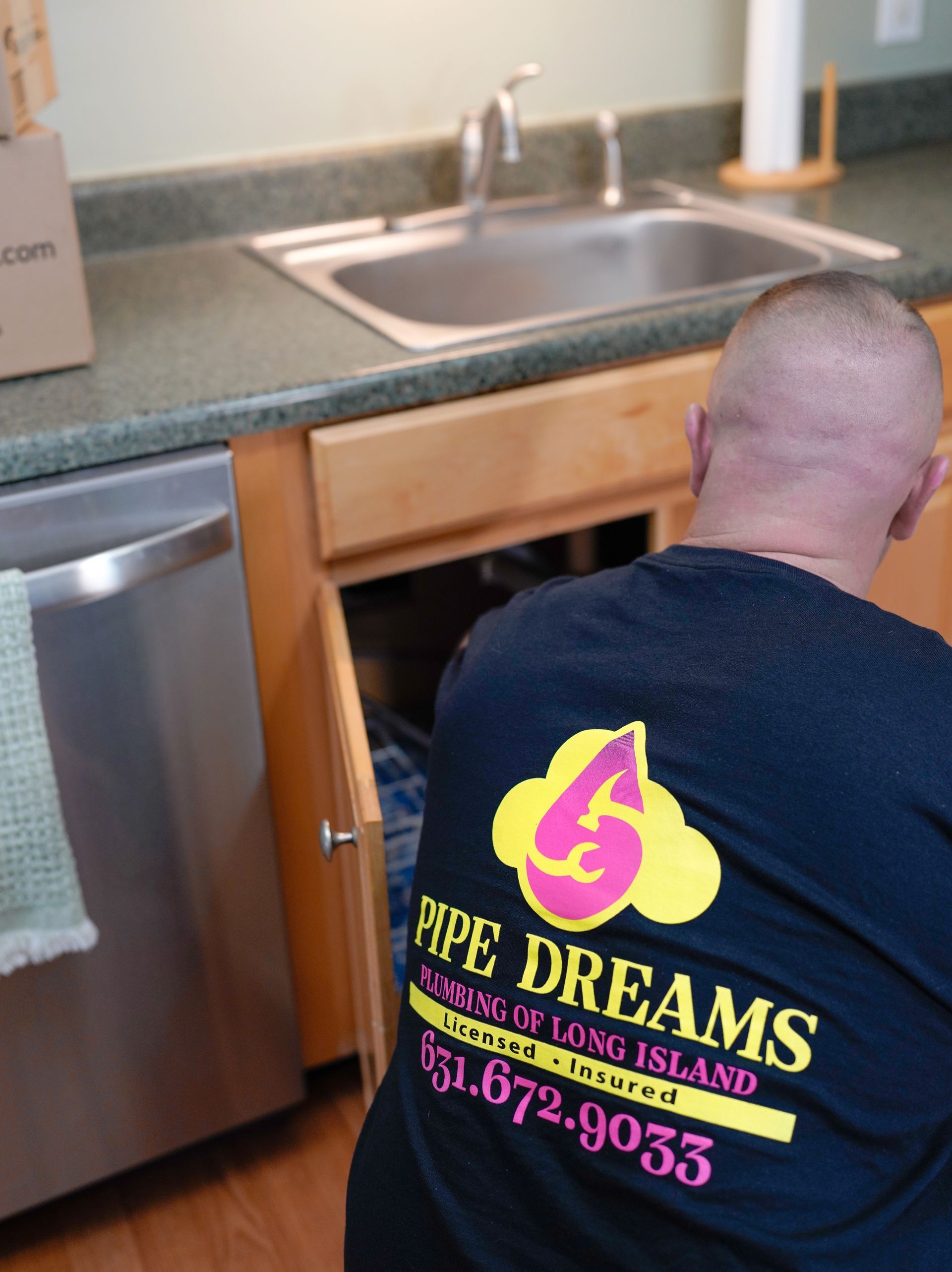 A plumber with a Pipe Dreams logo on their shirt works under a kitchen sink.