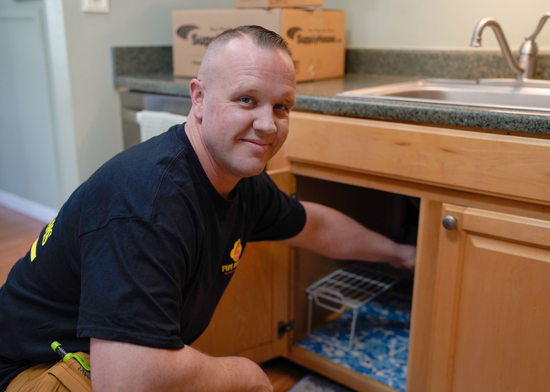 A person in a black t-shirt kneels to work under a wooden kitchen sink, looking toward the camera.