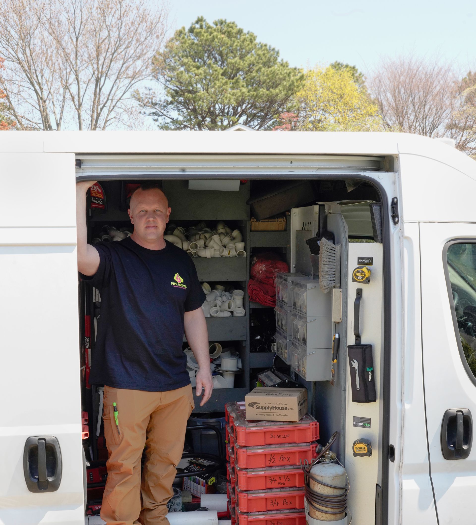 A technician stands in the open sliding door of a white service van filled with organized supplies, tools, and equipment.
