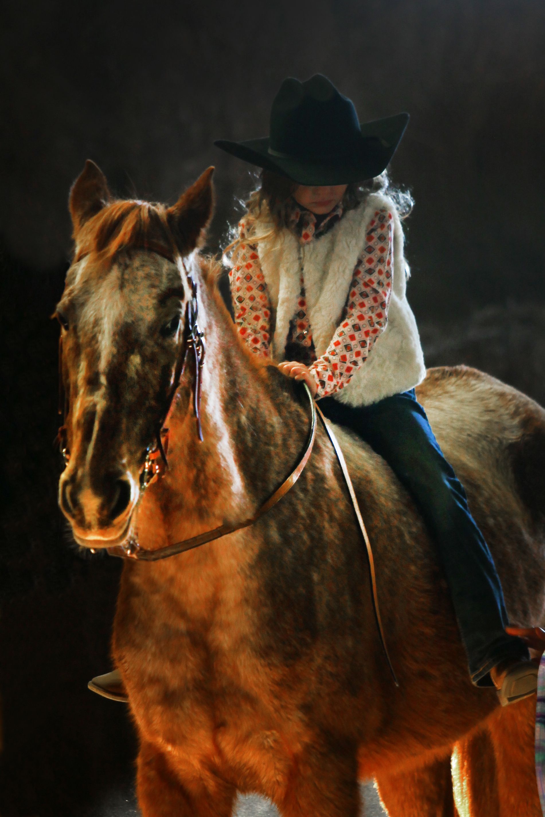 A little girl in a cowboy hat is riding a brown horse with dramatic lighting.