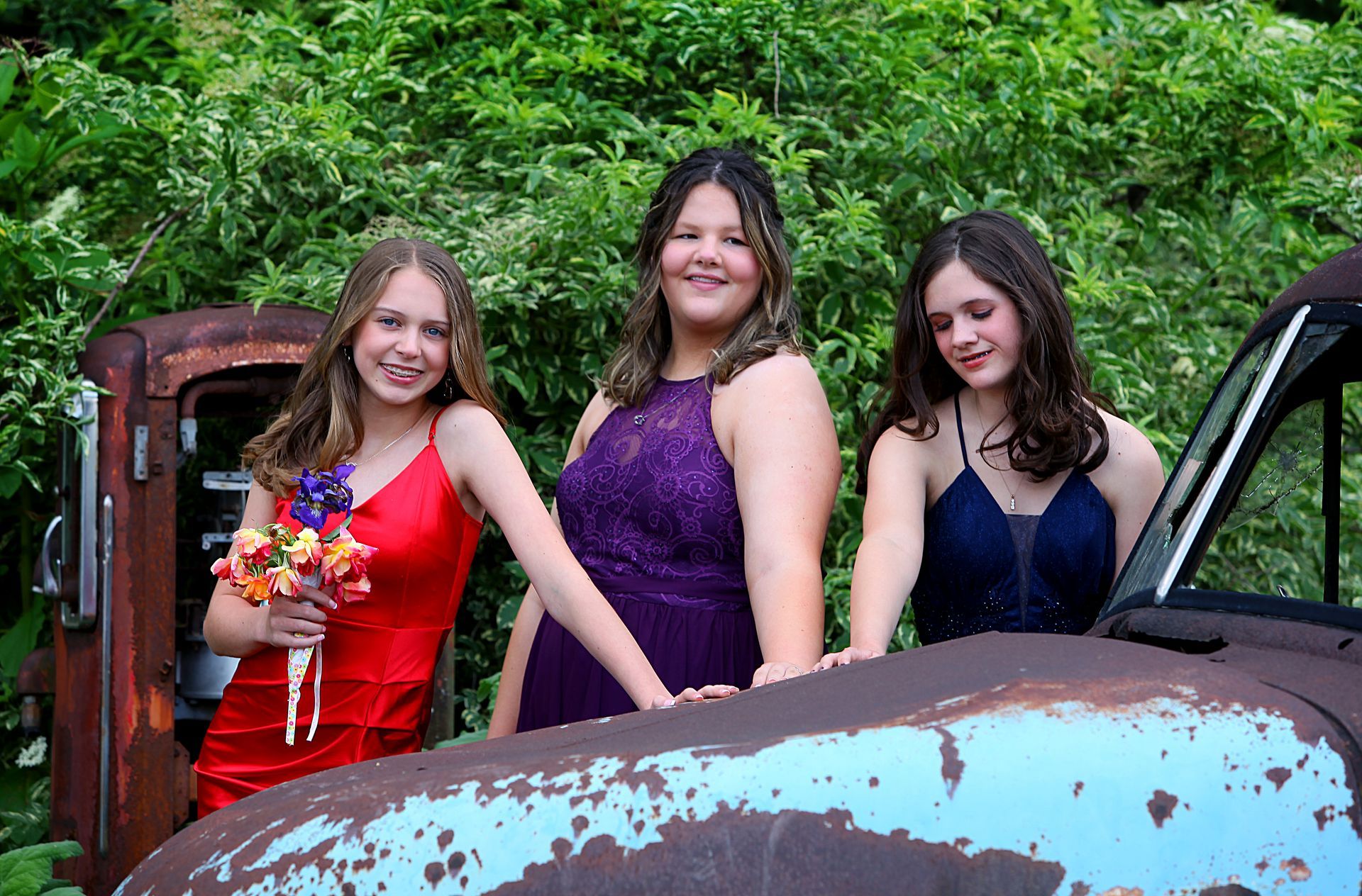 Three girls in dresses are posing for a picture in front of an old truck.