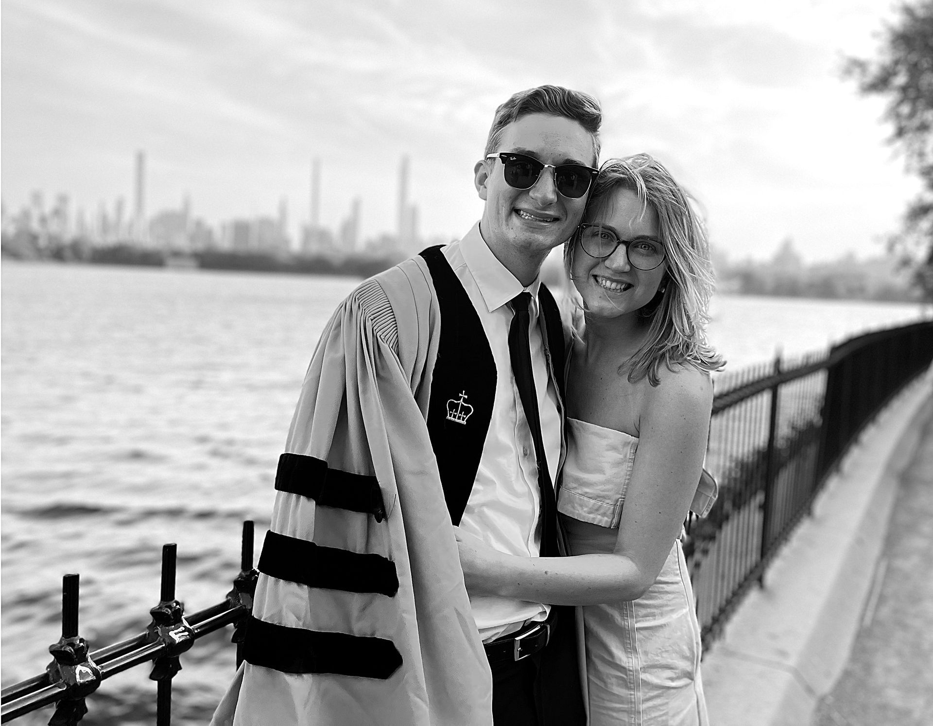 A man in graduation robes and a woman are posing for a picture in front of a lake with a cityscape in the background.