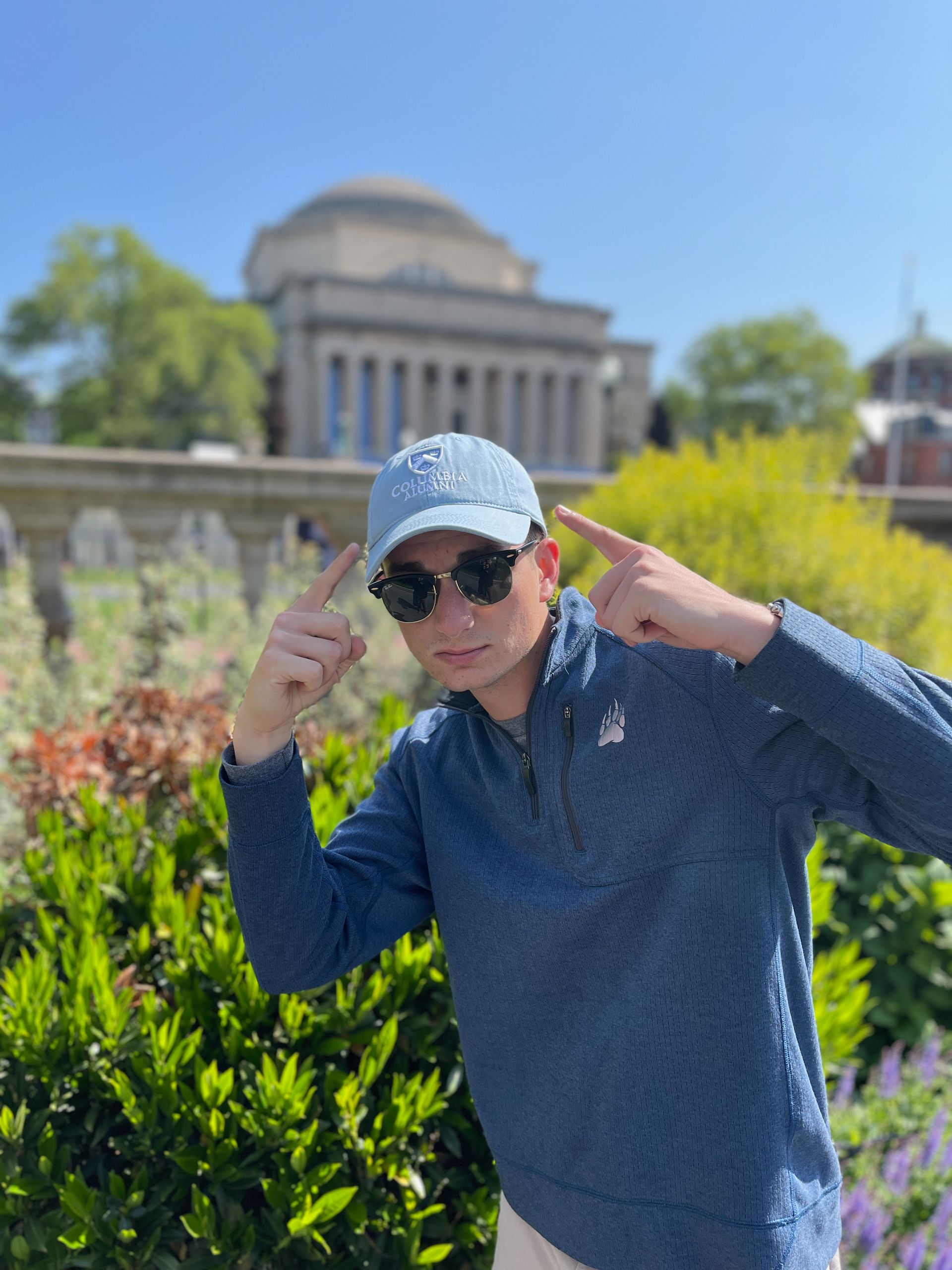 A man wearing sunglasses is pointing at his cap standing in front of a building a domed building.