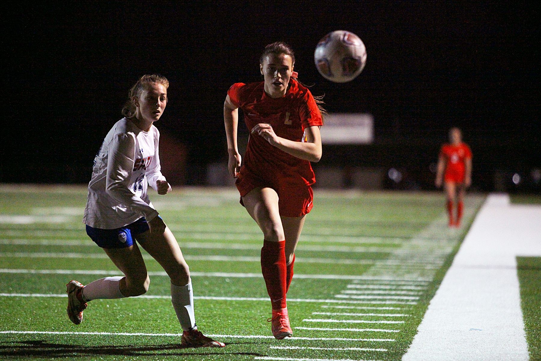 Two girls are playing soccer on a field at night.