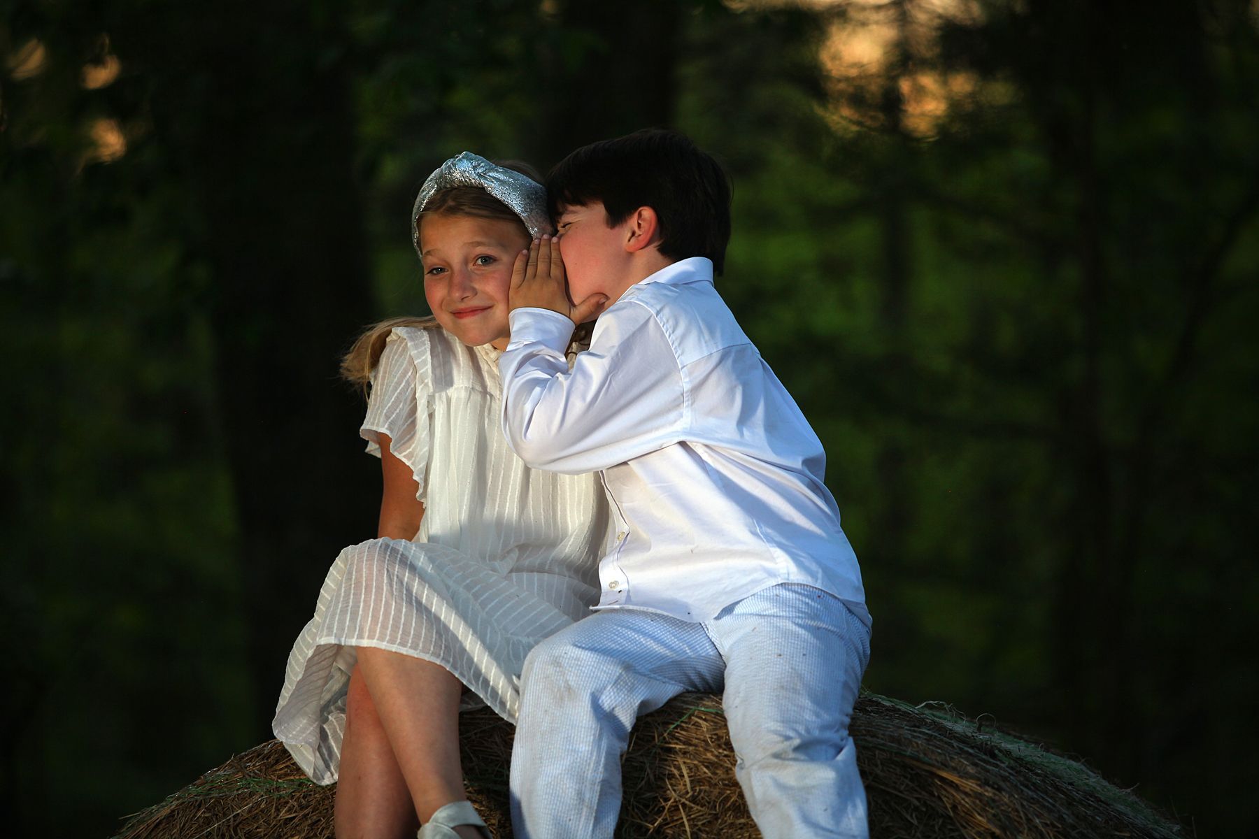 A boy is whispering into a girl 's ear while sitting on a bale of hay.