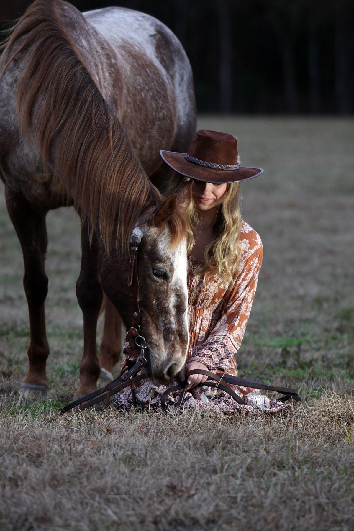 A woman in a cowboy hat is kneeling next to a horse in a field.