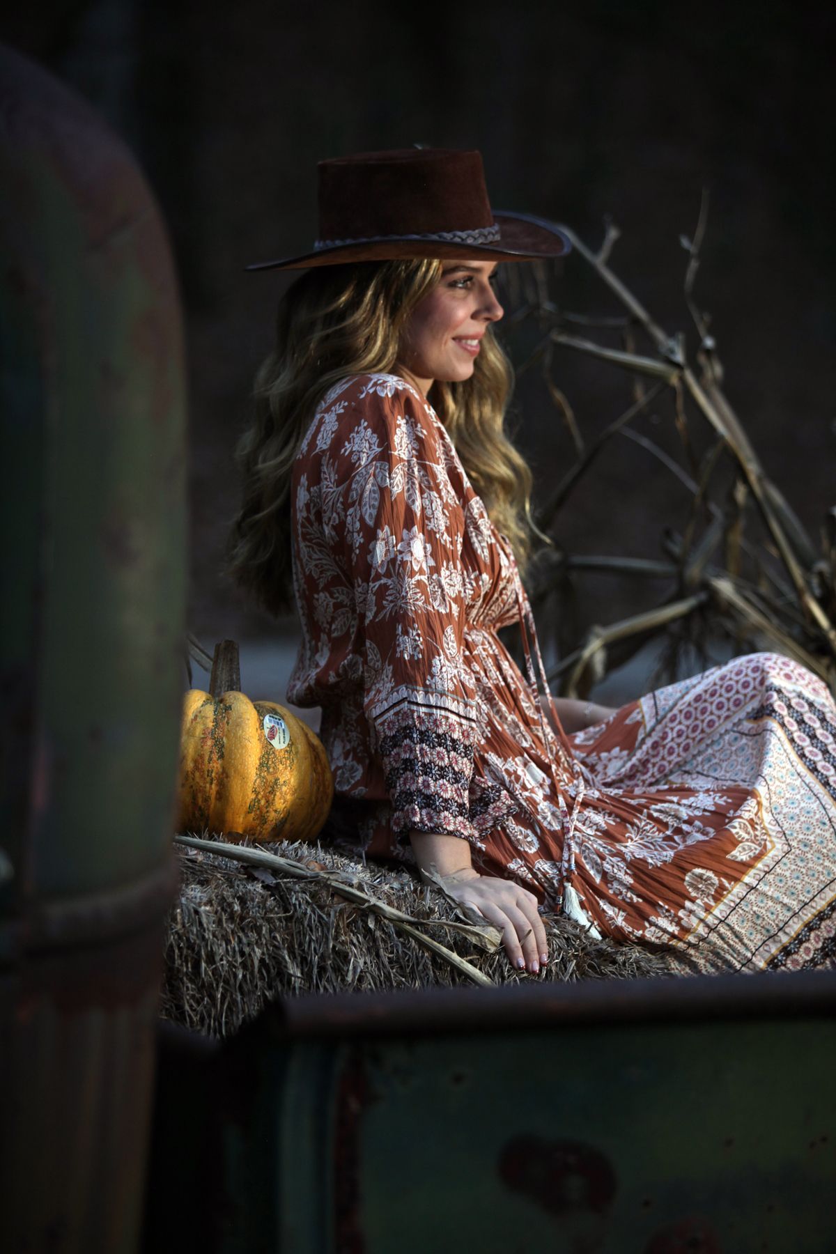 A woman in a hat is sitting on a hay bale next to a pumpkin.