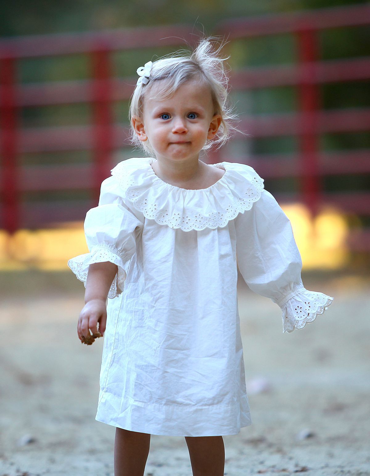 A little girl in a white dress is standing in front of a red fence.