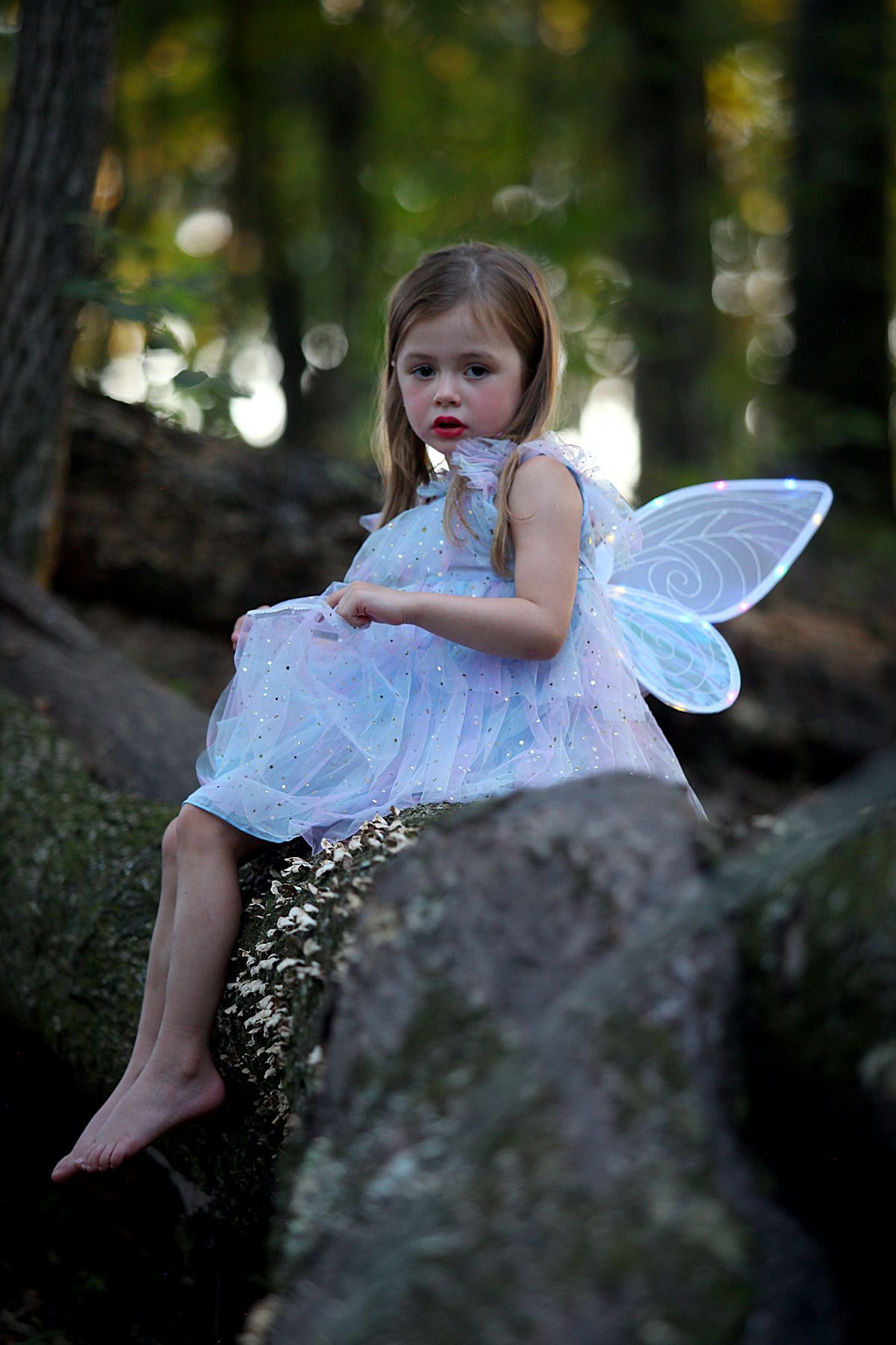 A little girl dressed as a fairy is sitting on a rock in the woods.