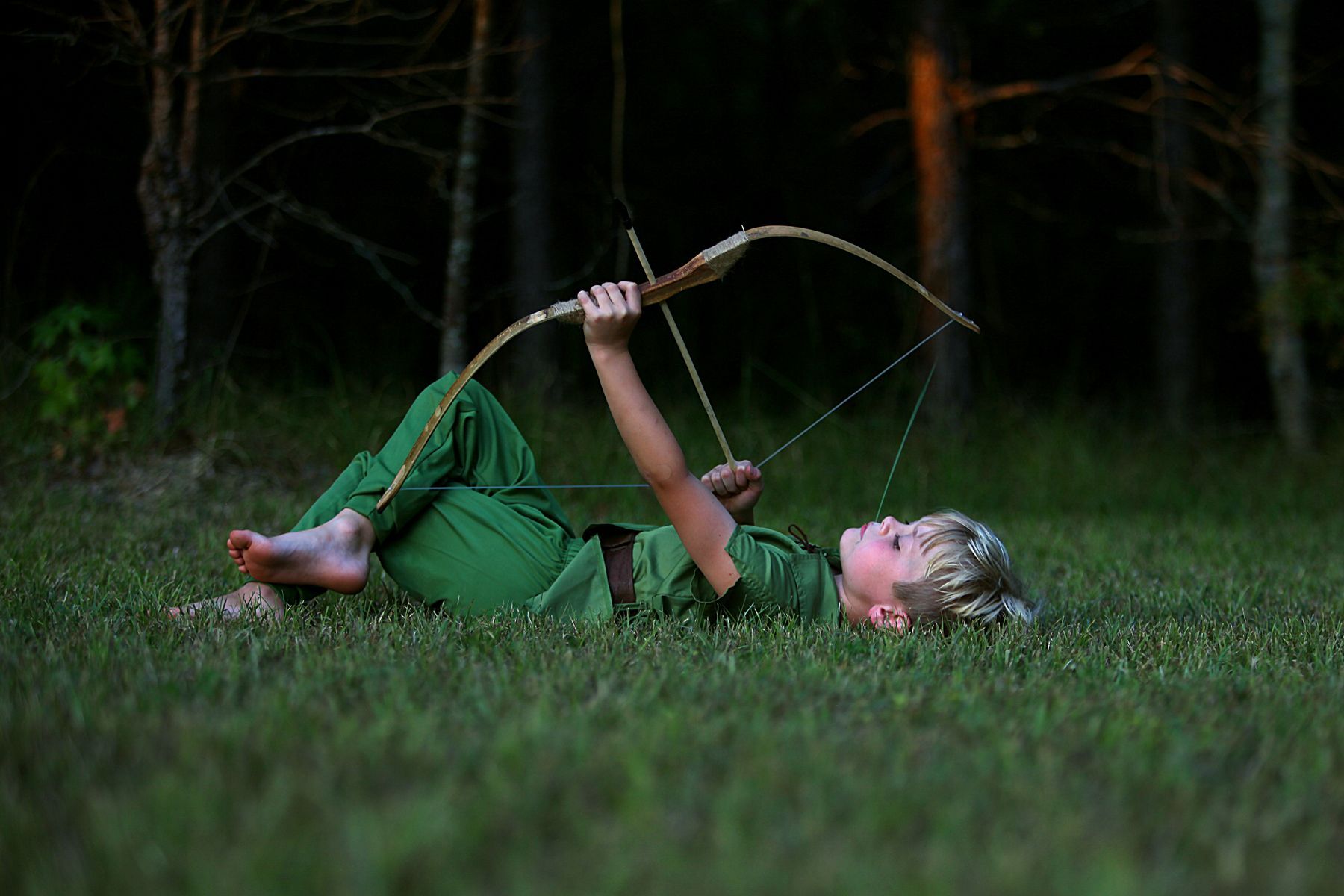 A little boy dressed as Peter Pan is lying in the grass holding a bow and arrow.