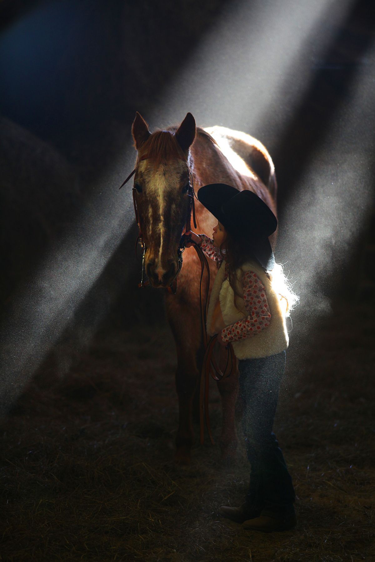 A little girl in a cowboy hat is standing next to a horse in a dark room.