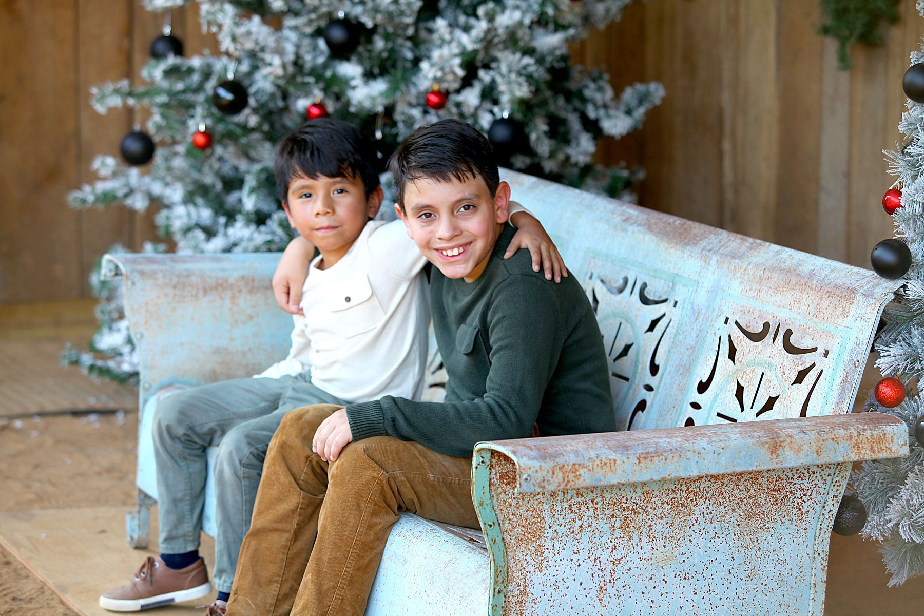 Two young boys are sitting on a bench in front of a christmas tree.