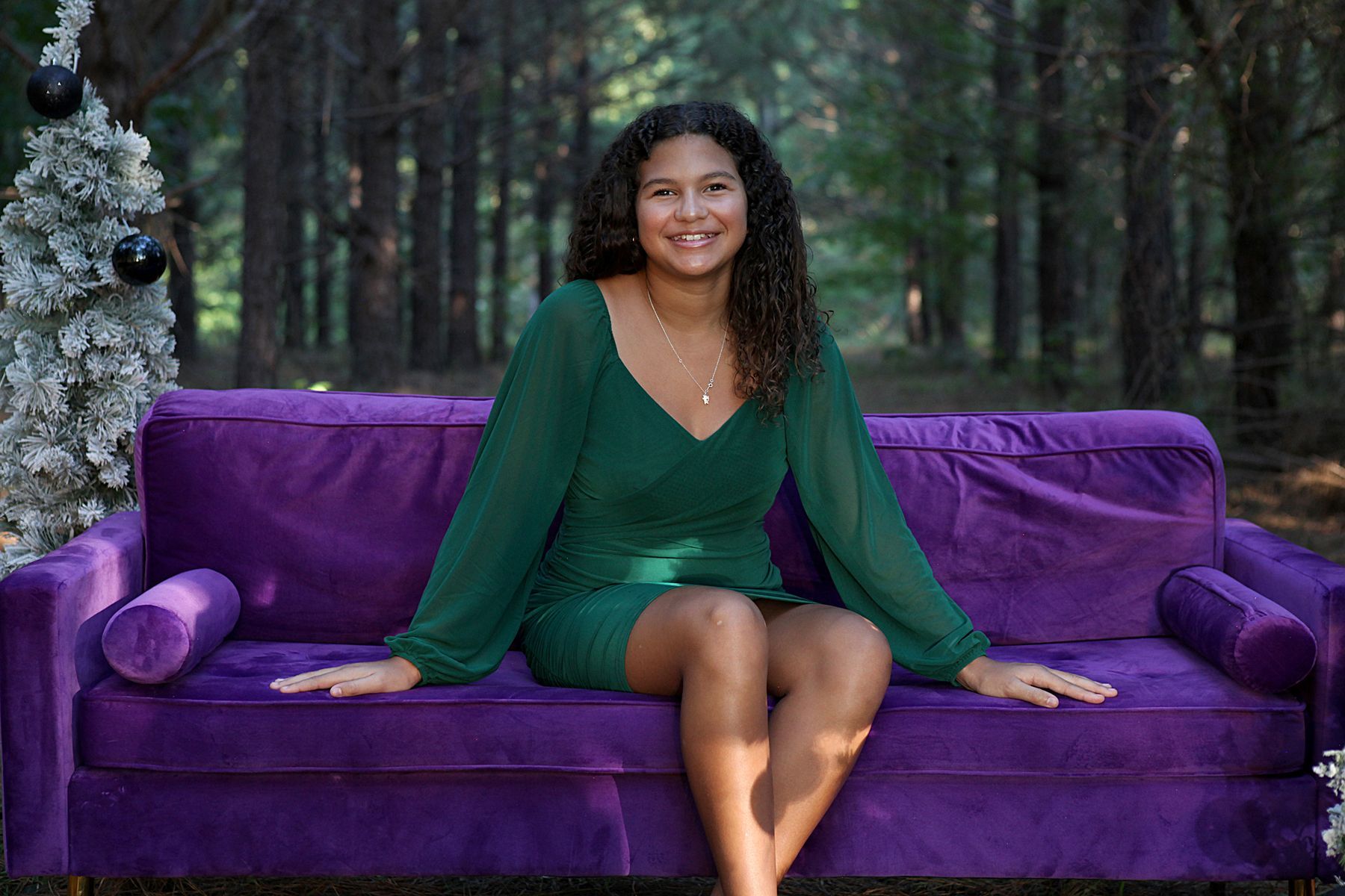 A woman in a green dress is sitting on a purple couch in the woods.