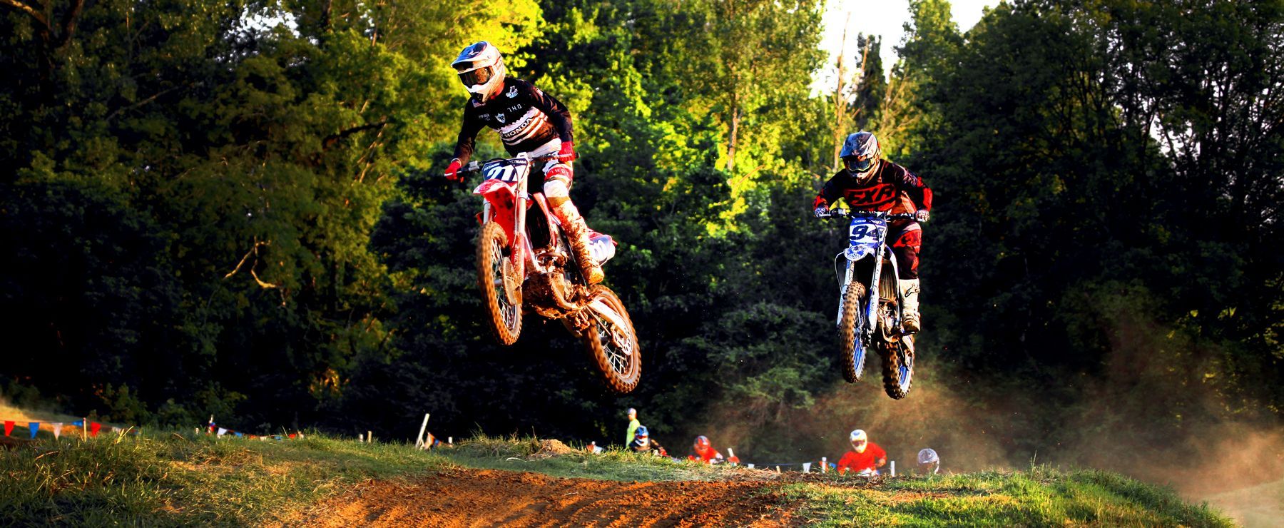 Two motocross riders are jumping in the air on a dirt track.