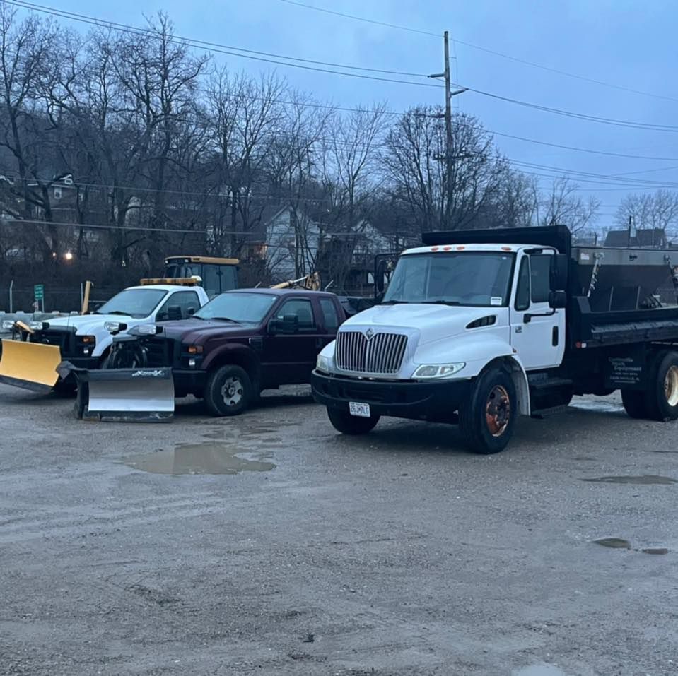 Three snow removal trucks parked outdoors. One white dump truck and two with plows. Trees in the background.