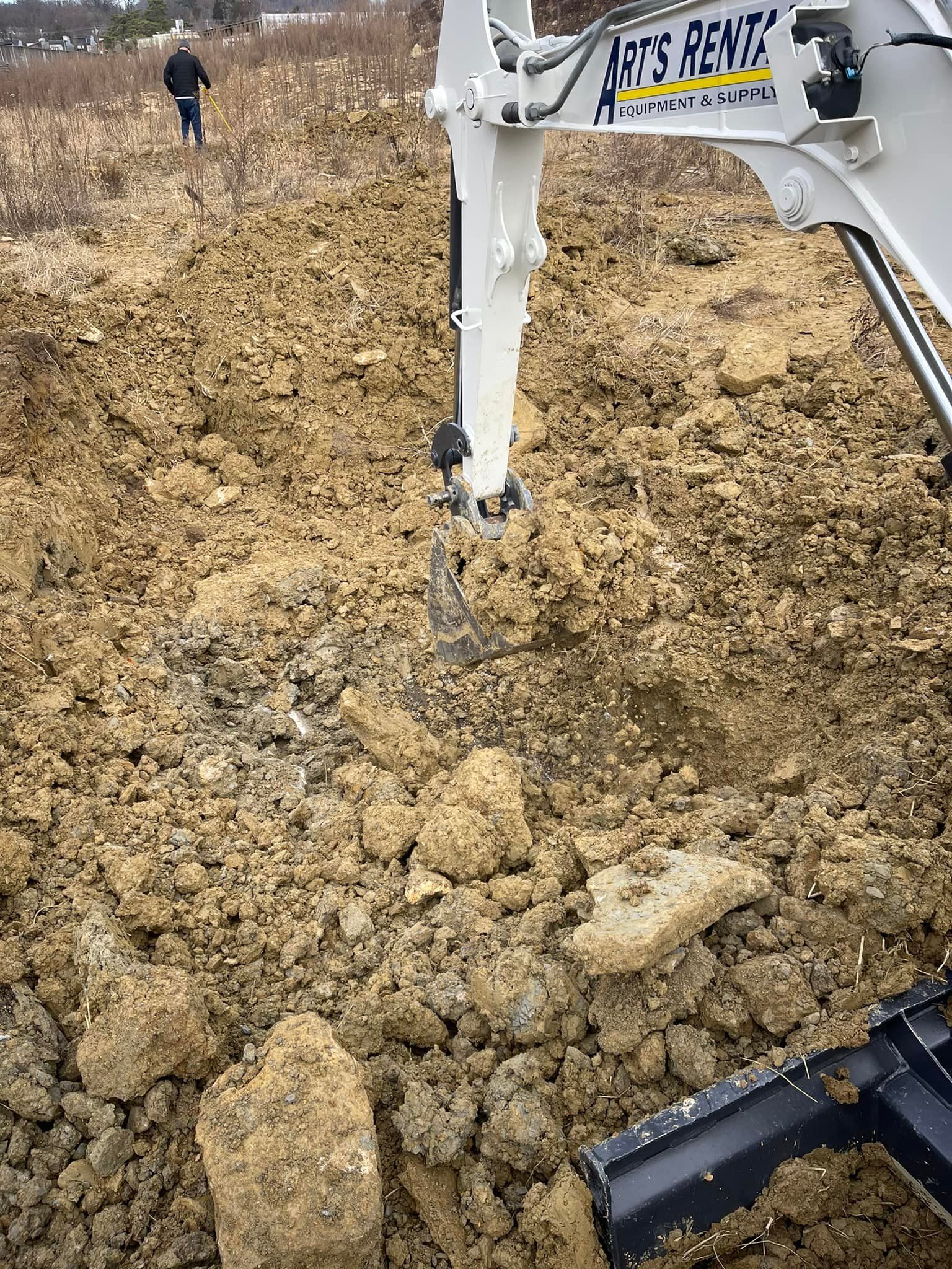 Excavator digging in dirt; a person walks in the background. Cloudy sky.