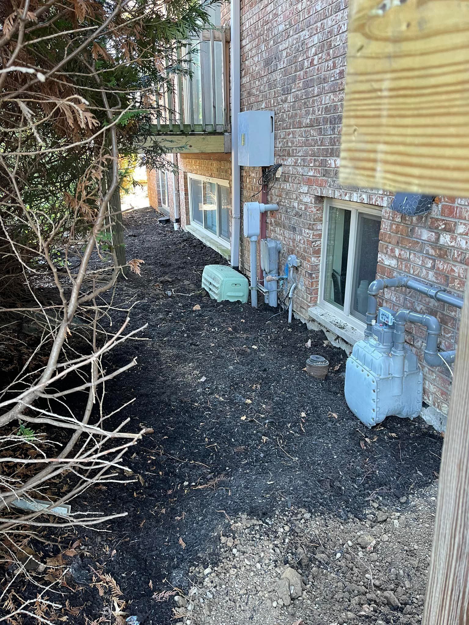 Dark mulch bed along a brick building, with windows and a balcony. A propane tank and electrical boxes are visible.