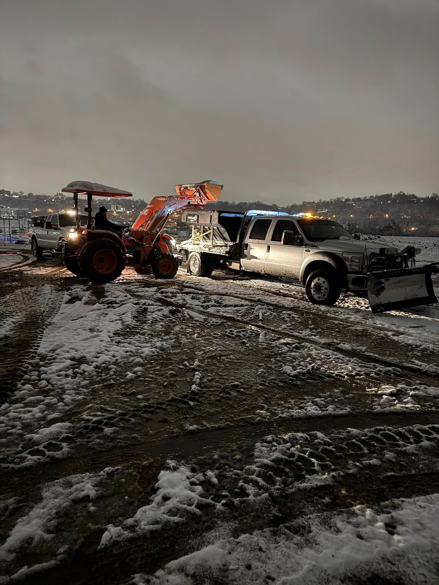 Snow-covered parking lot with a pickup truck and tractor clearing snow at night.