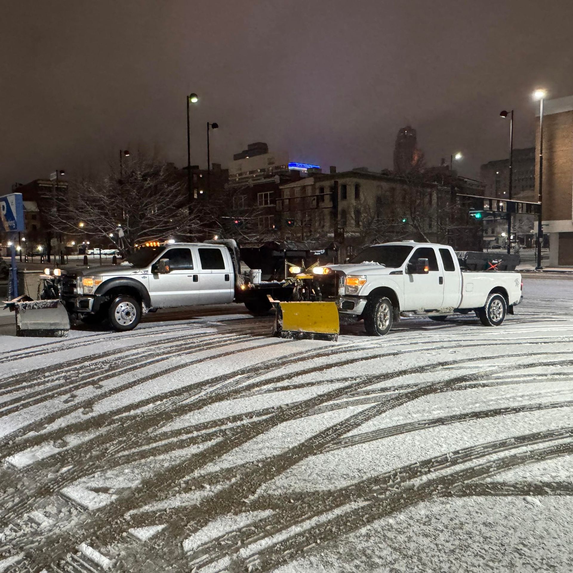 Two snow plows clearing a snowy urban area at night. One silver, one white. Buildings in the background.