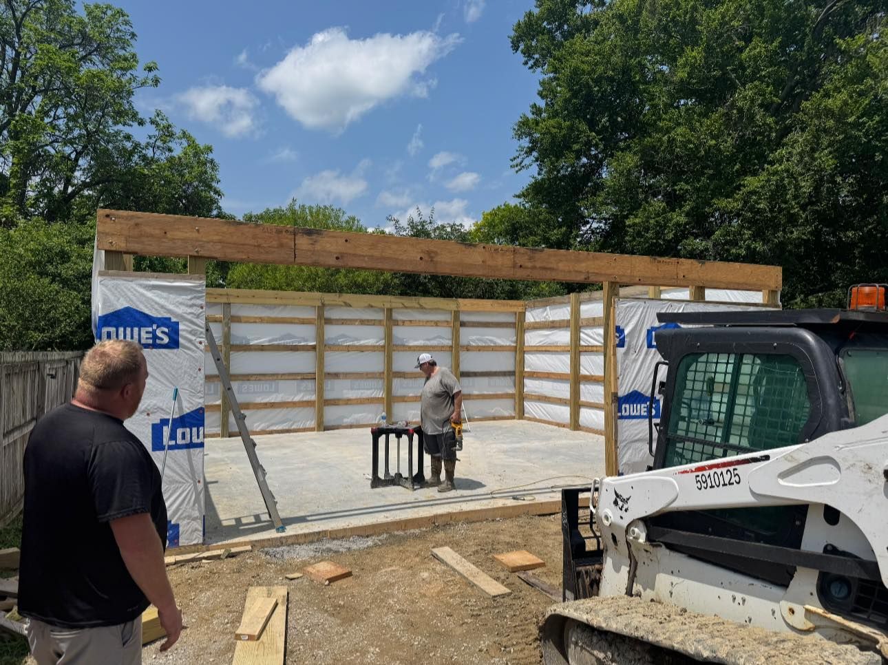Construction of a garage: two men and a skid steer. Wooden frame, blue skies, concrete floor, Lowe's wrap.