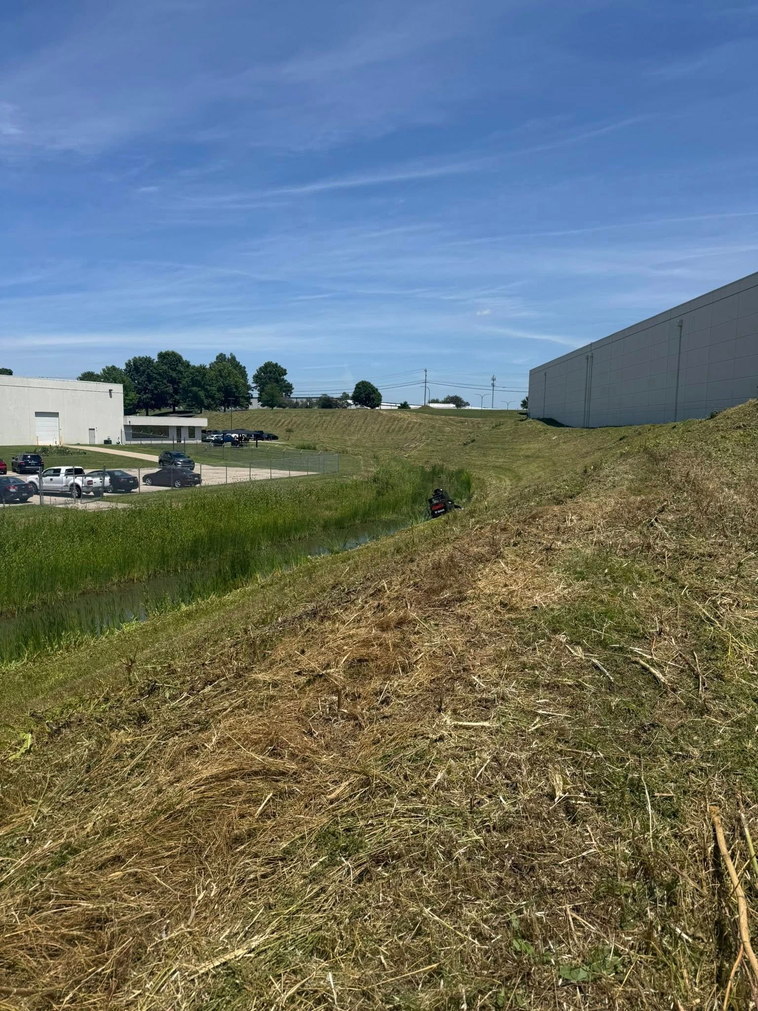 Pile of wood chips and vegetation in front of industrial buildings, under a blue sky.
