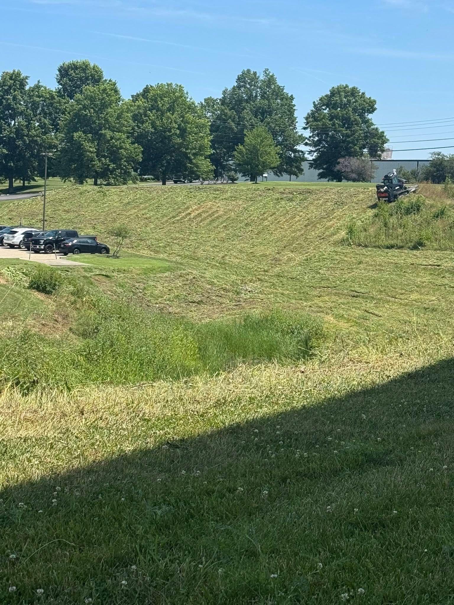 A person operating a lawn mower on a grassy hillside under a blue sky, with trees in the background.