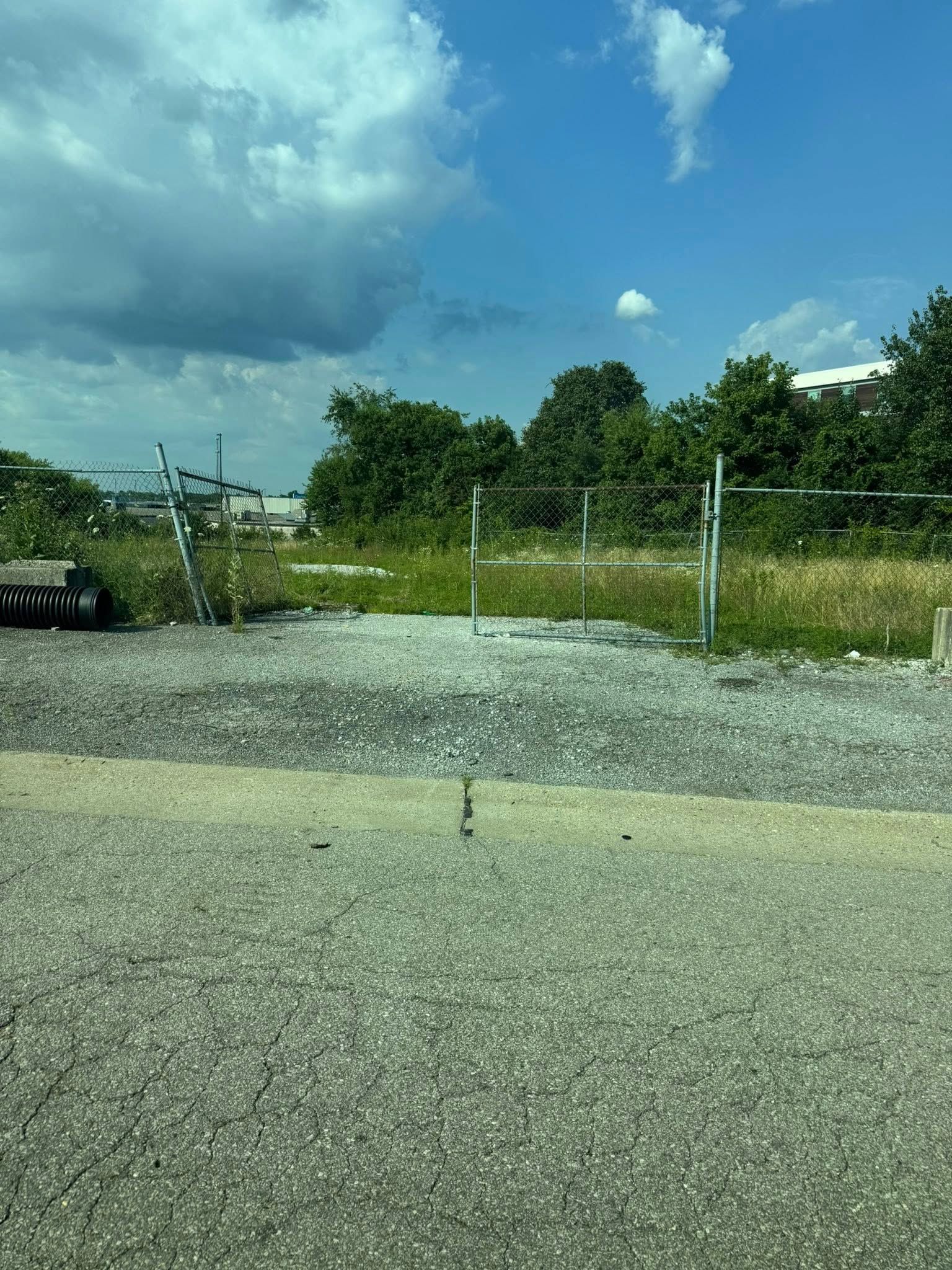 Gravel lot with metal fence, overgrown grass, and trees under a cloudy blue sky.