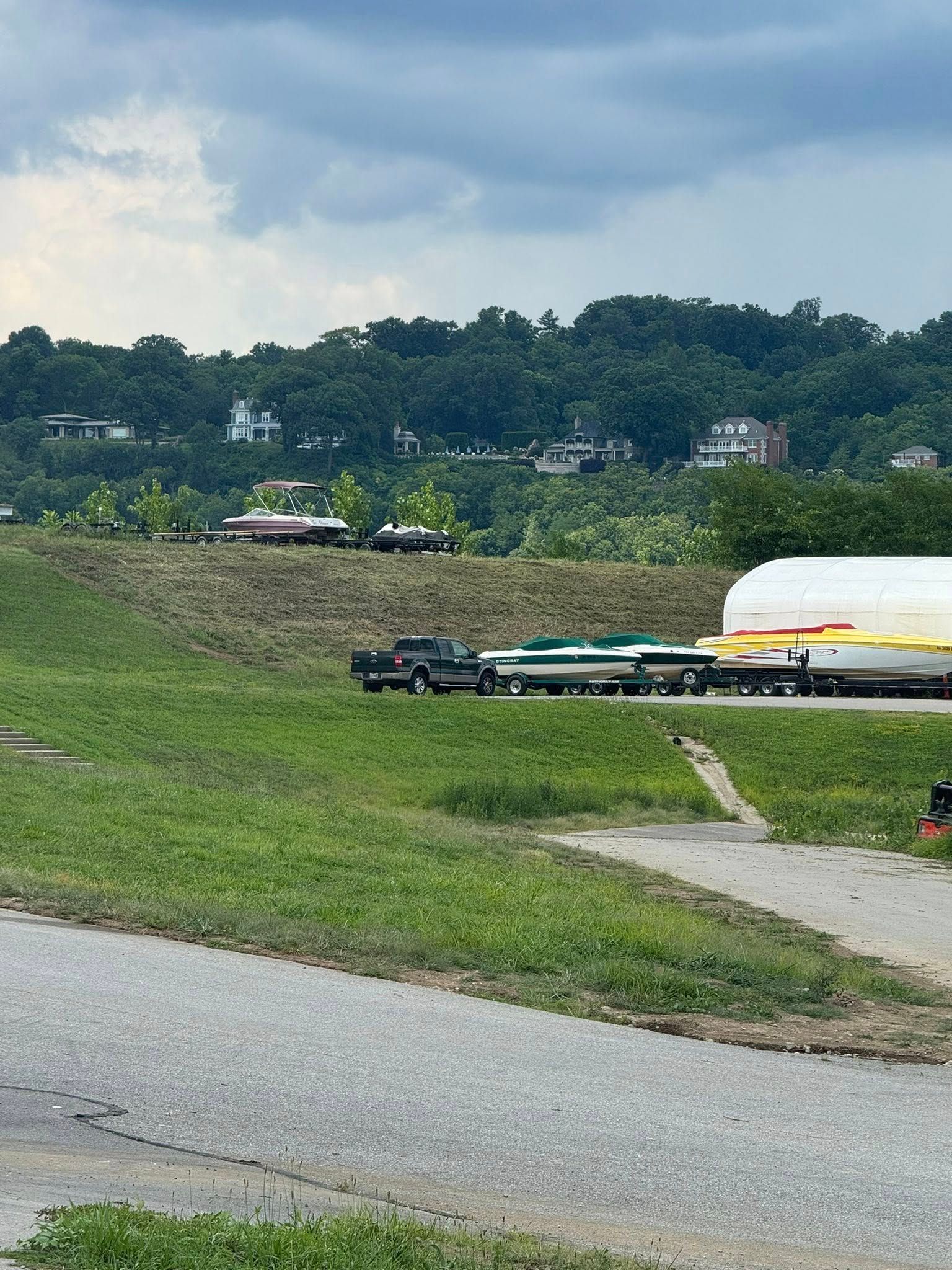 Truck hauling several covered boats on a grassy field, houses on a hillside in the background.