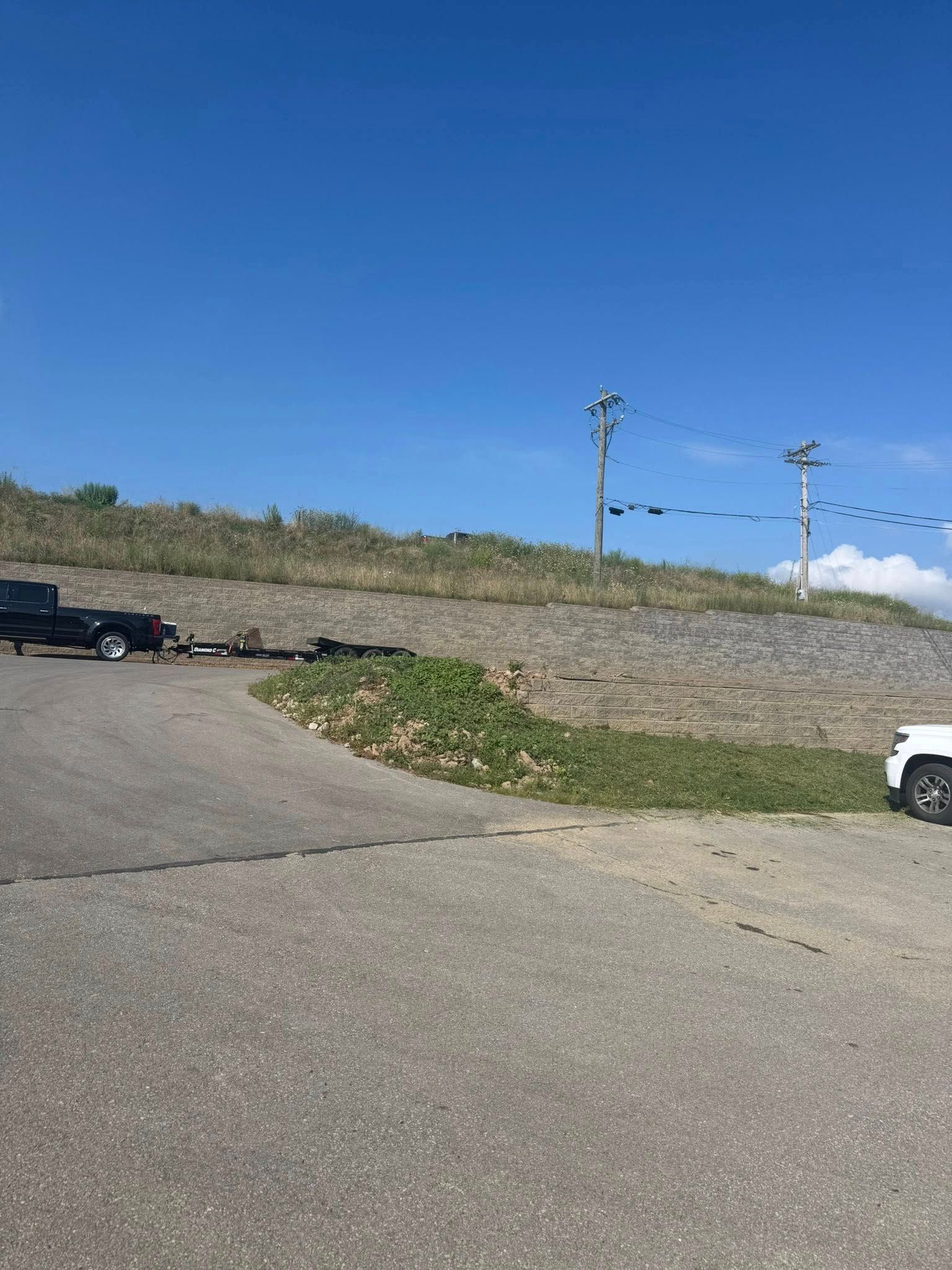 Paved road curves with a truck and trailer, and another vehicle parked on the right. Electric poles against a blue sky.
