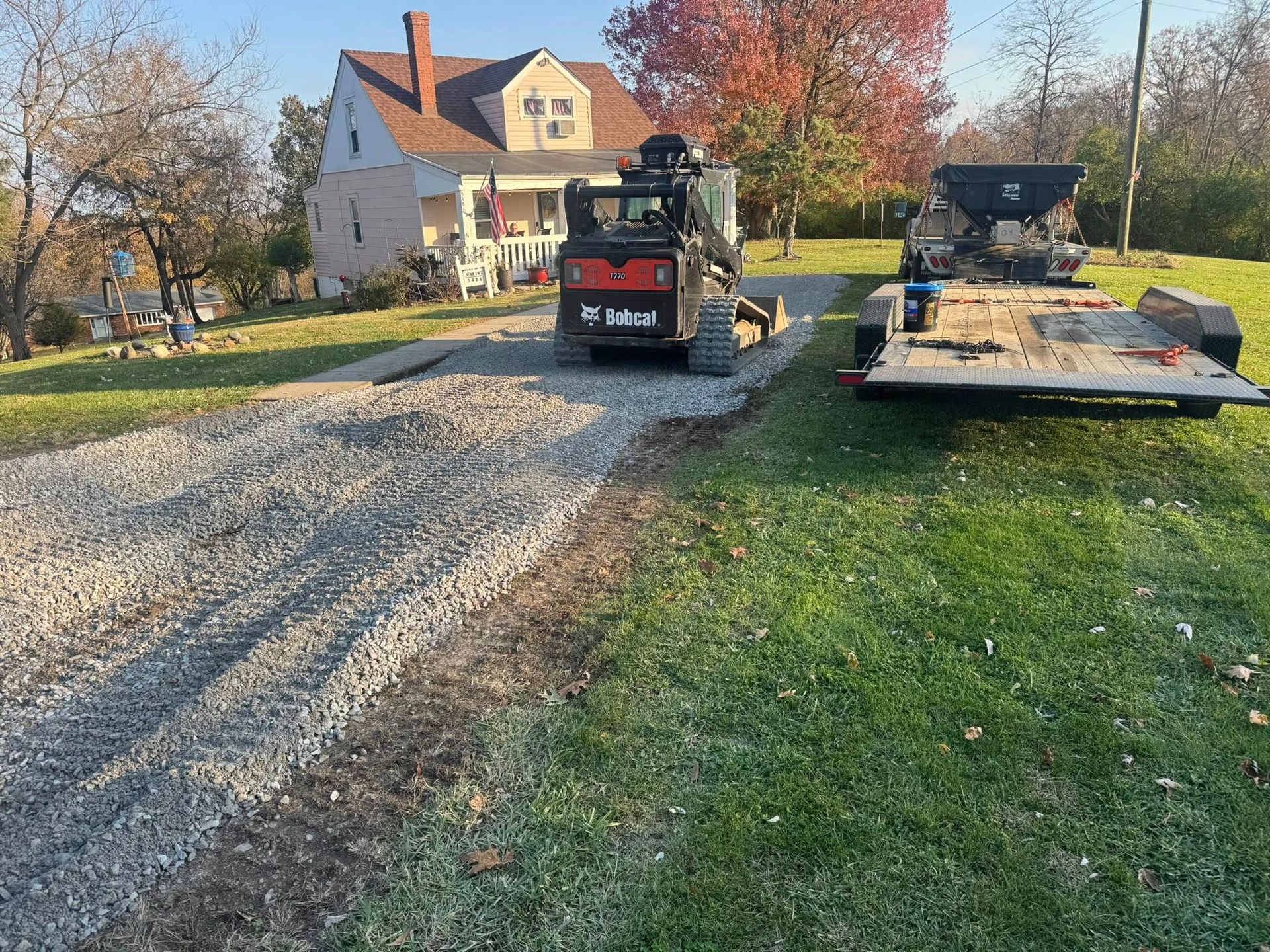 Gravel driveway under construction, with a Bobcat and trailer on a grassy lot next to a house.