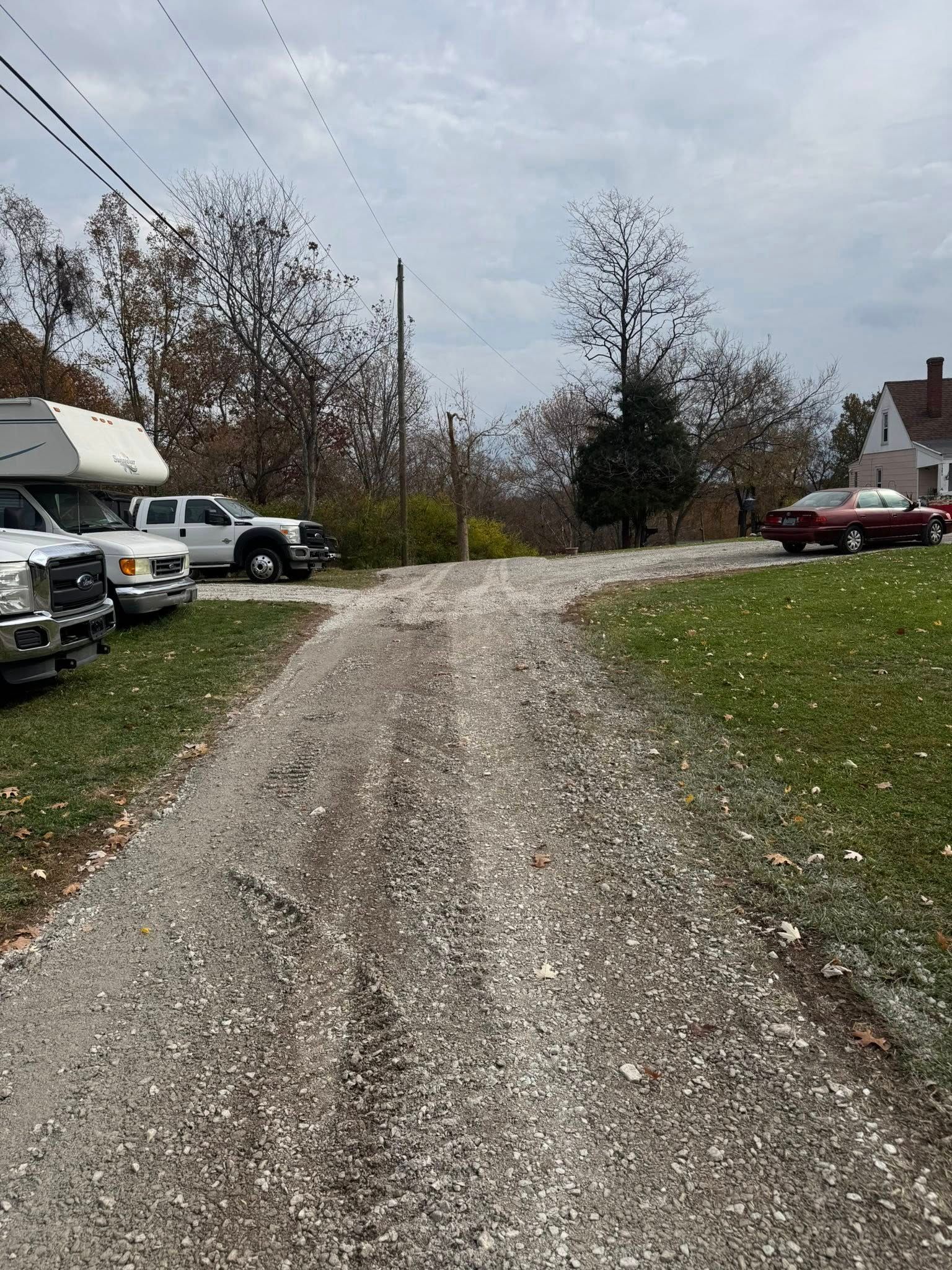 A gravel driveway splits, leading past parked trucks and a red car on a lawn under a cloudy sky.