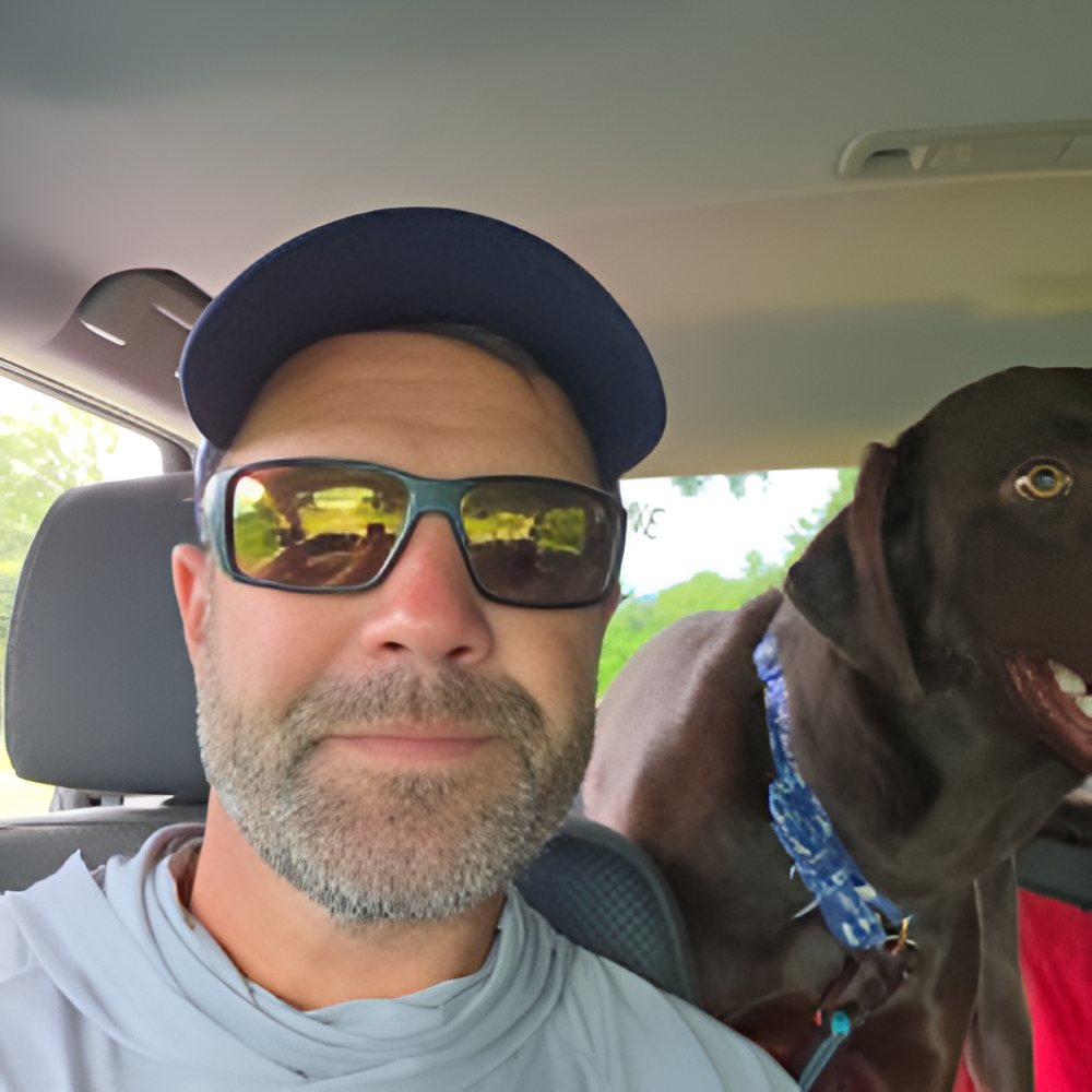 Man in sunglasses and hat smiles with a brown dog in a car.