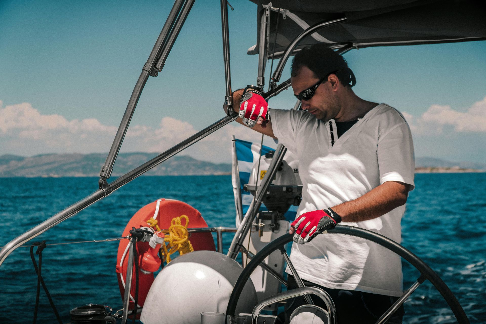 Man in sunglasses steers sailboat on sunny sea; red gloves, Greek flag.