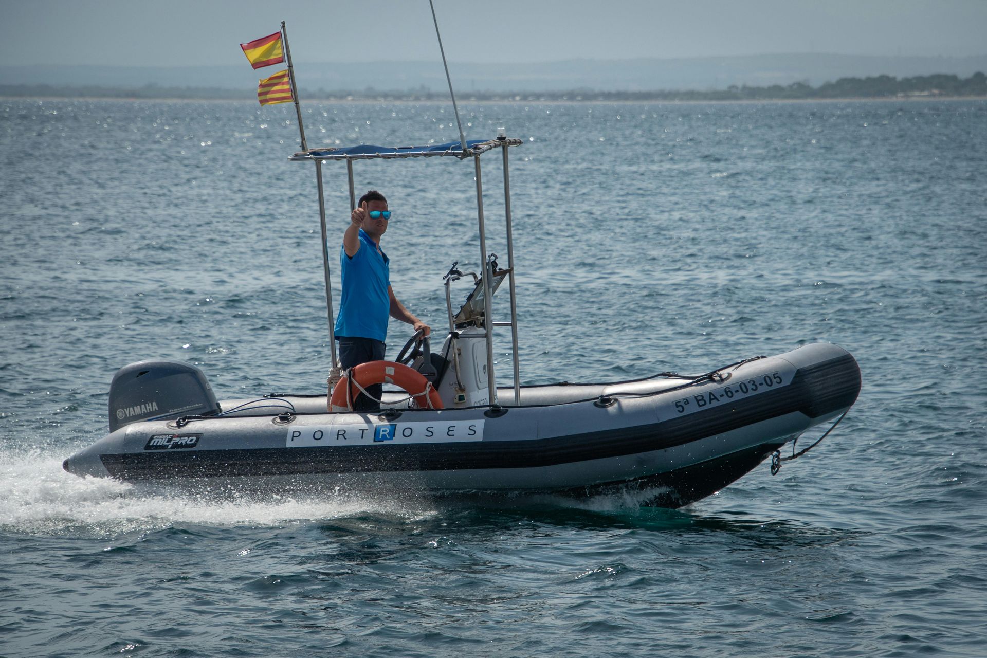 Man waves from a gray inflatable boat, driving on water with Spanish flag; sunny day.