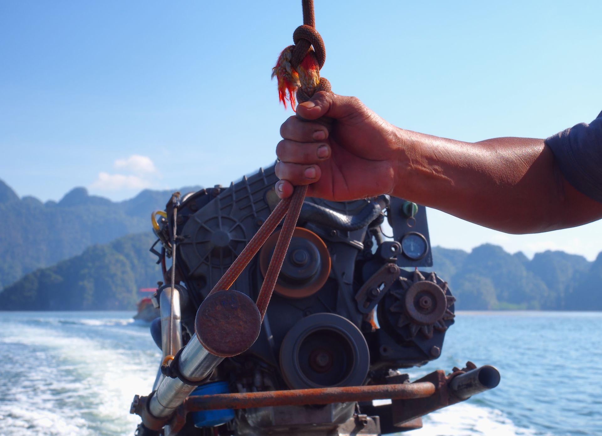 Hand holding a rope attached to a boat engine, over water, with mountains in the distance.