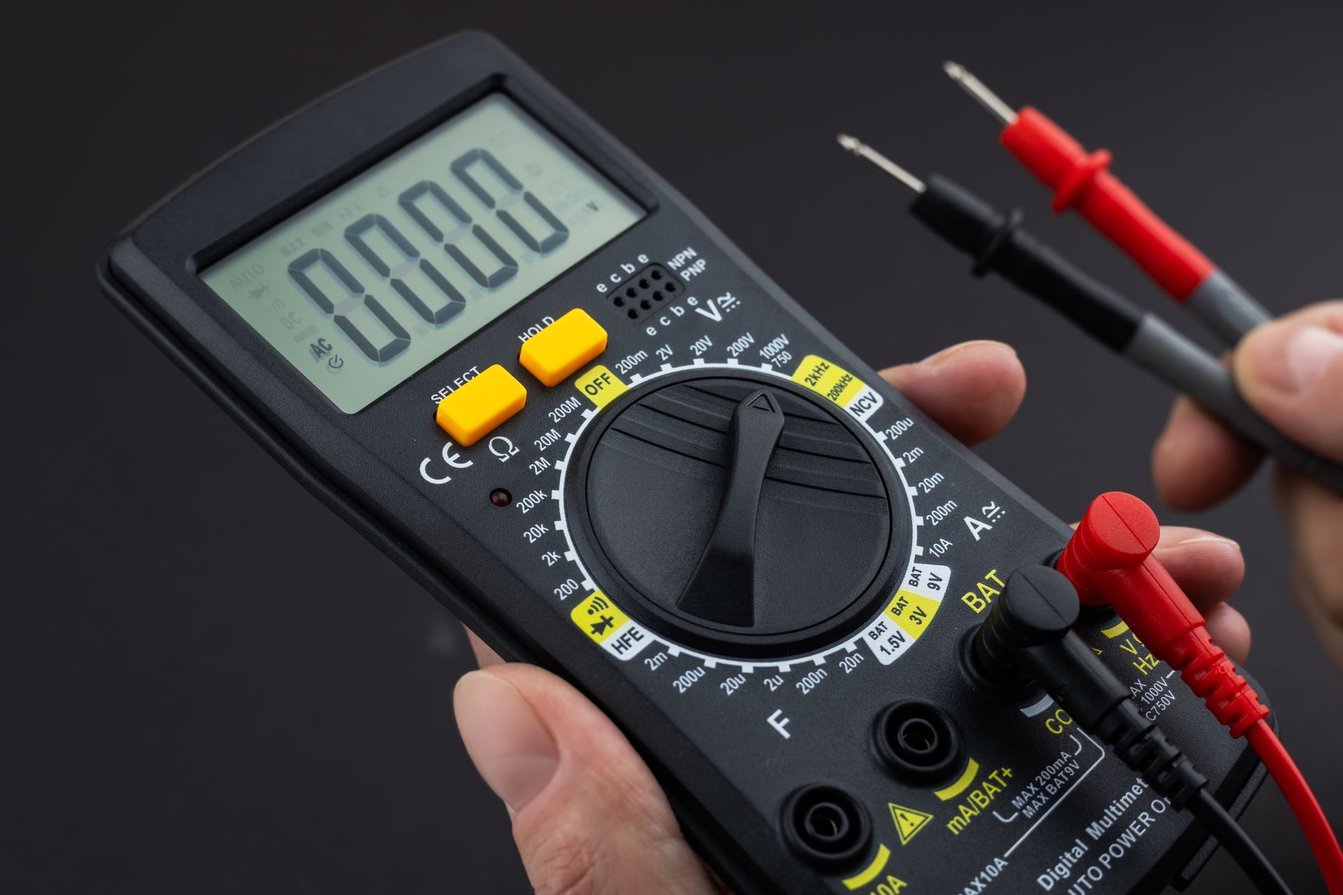 Hand holding a digital multimeter with probes. The display reads 0000.