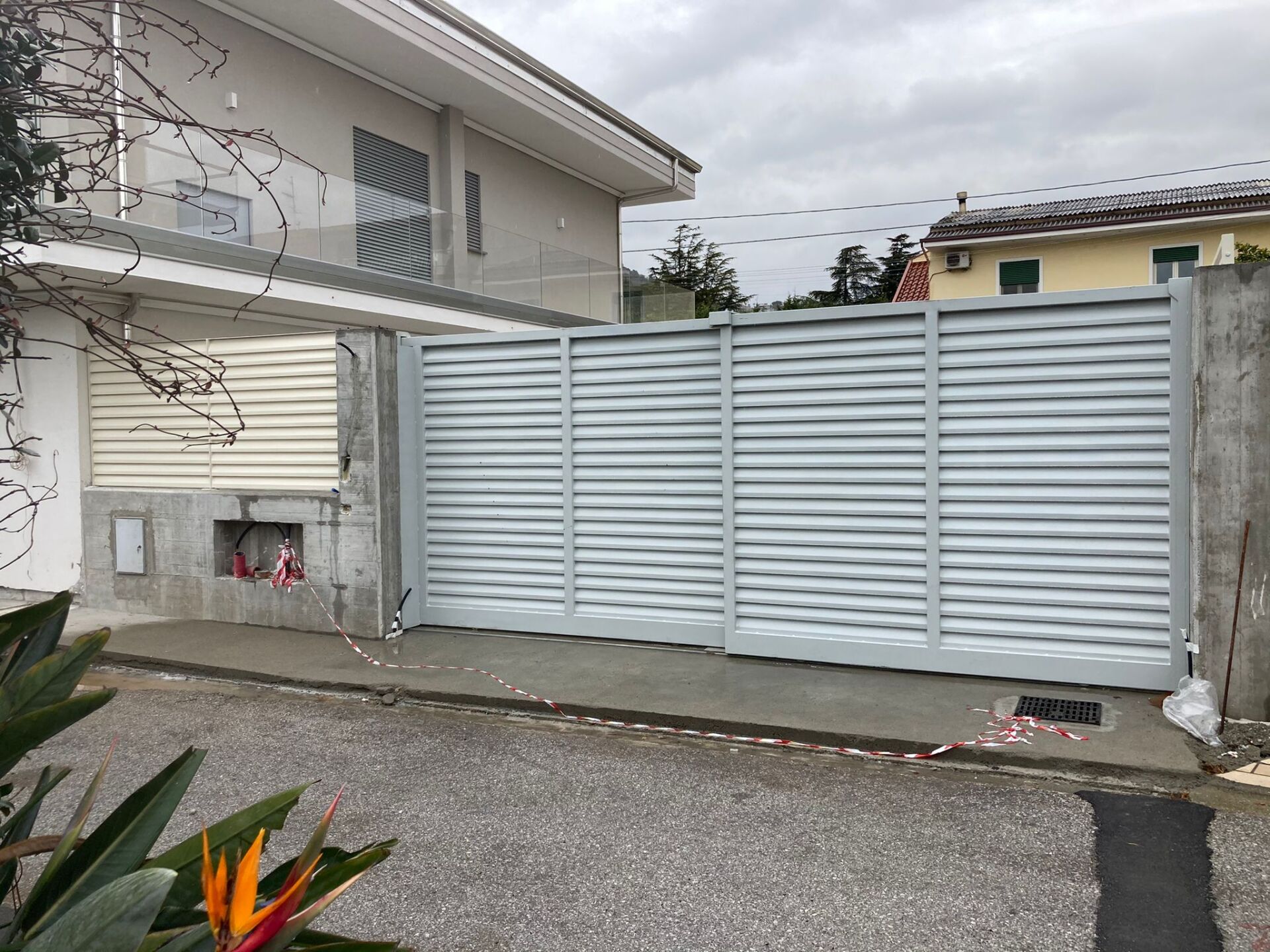 Cancello orizzontale grigio chiaro, muri di cemento, casa sullo sfondo, giornata nuvolosa.