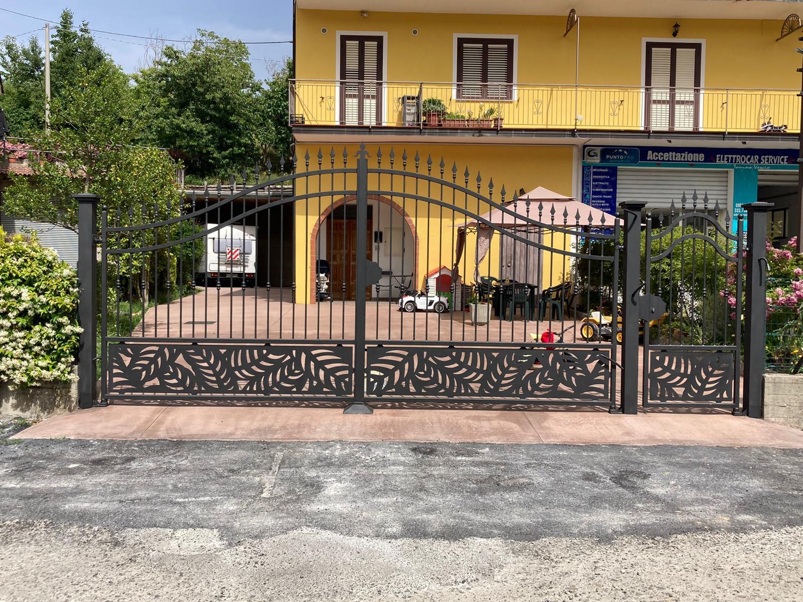 Cancello carrabile in metallo nero con motivo a foglie; edificio giallo sullo sfondo.
