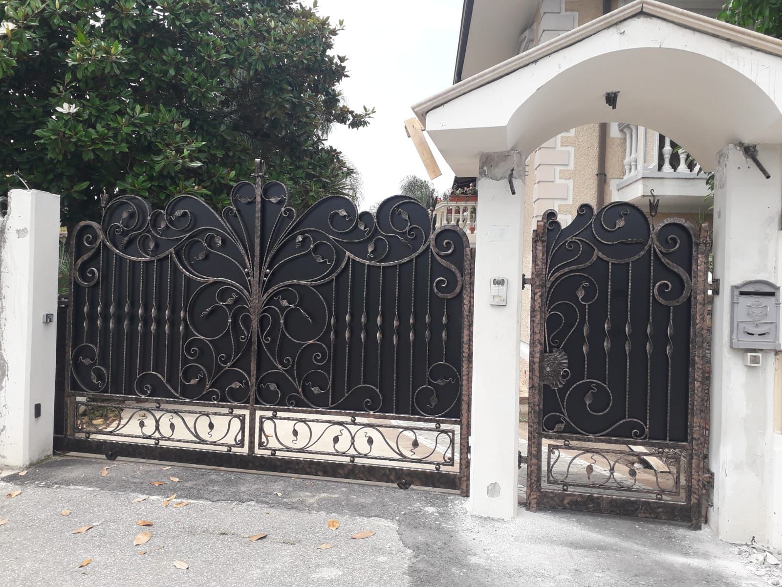 Cancello in ferro battuto nero e porta laterale con volute decorative, incastonati in un ingresso in pietra con cassetta della posta.