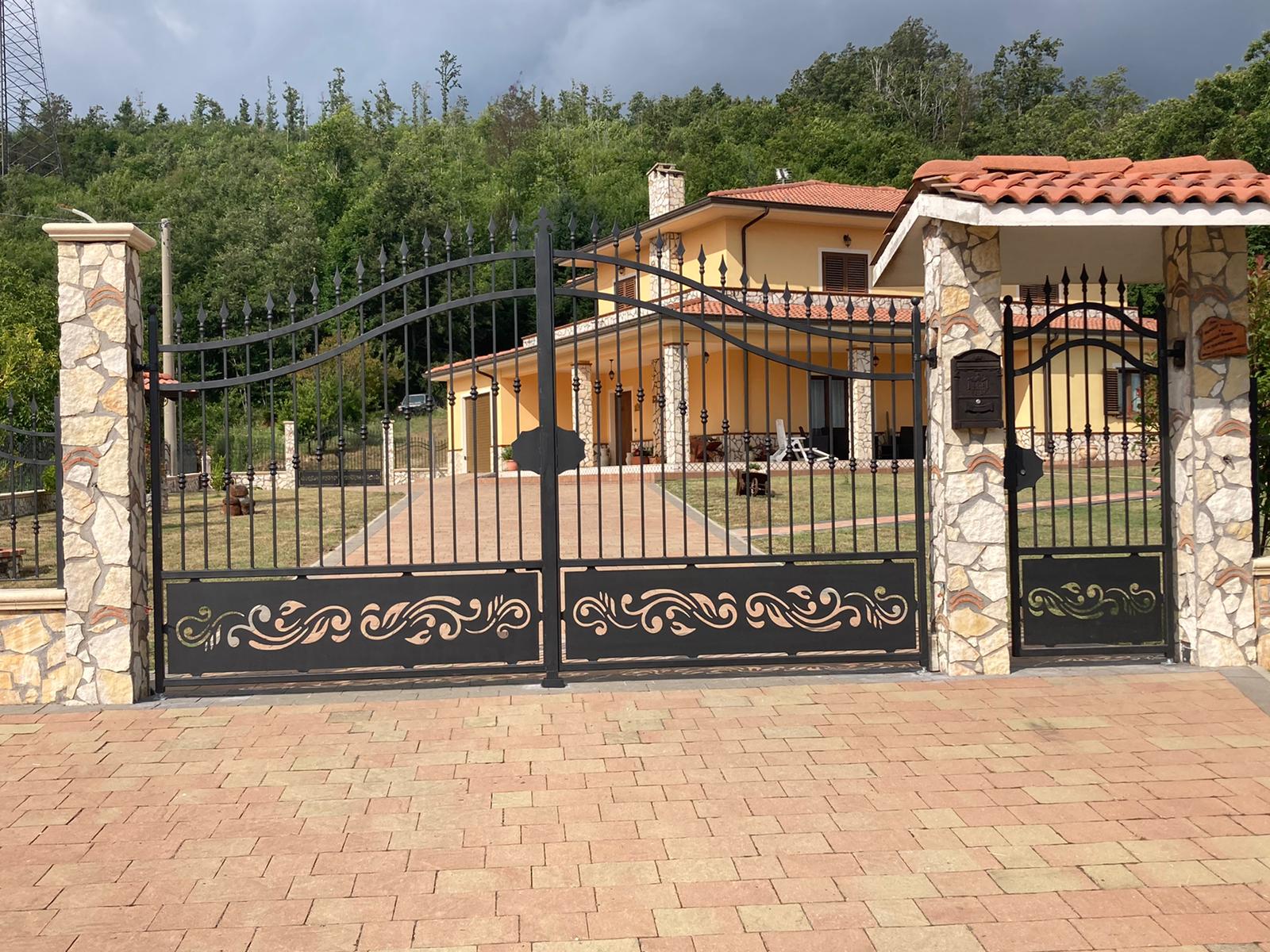 Cancelli in ferro battuto nero con motivi ornamentali conducono a una casa gialla con tetto in tegole rosse, vialetto in mattoni e pilastri in pietra.