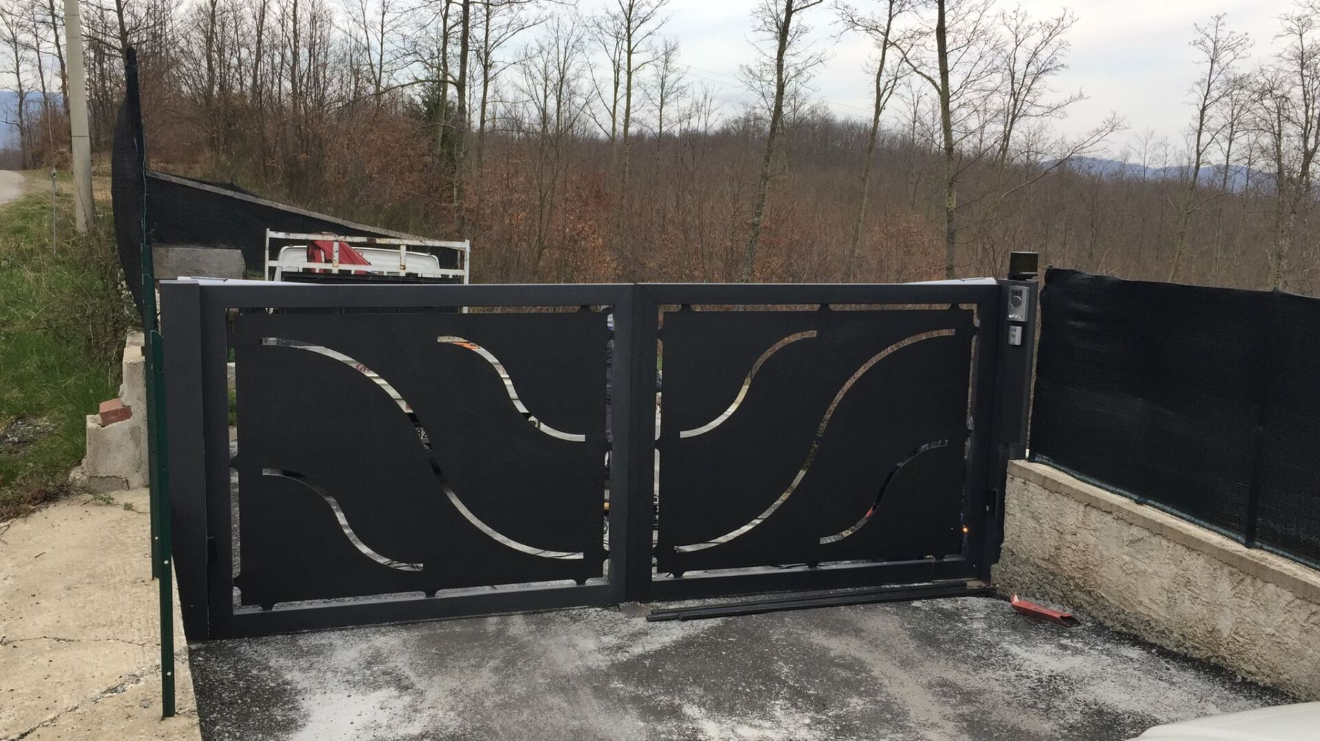 Cancello d'accesso in metallo nero con motivo a onde, incastonato tra una collina e degli alberi.